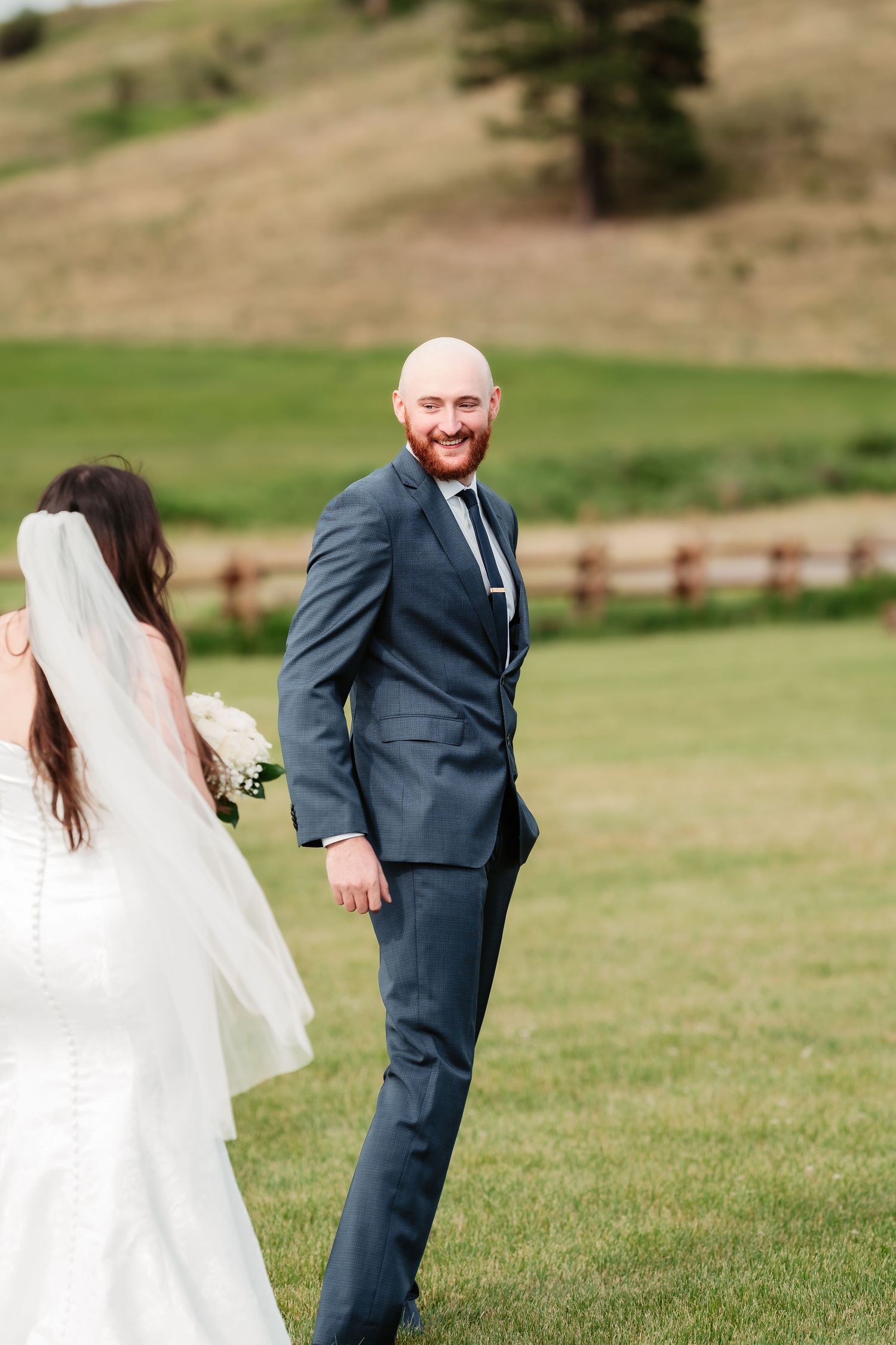 Wedding couple stands in a scenic mountain meadow with wooden fence and rolling hills.