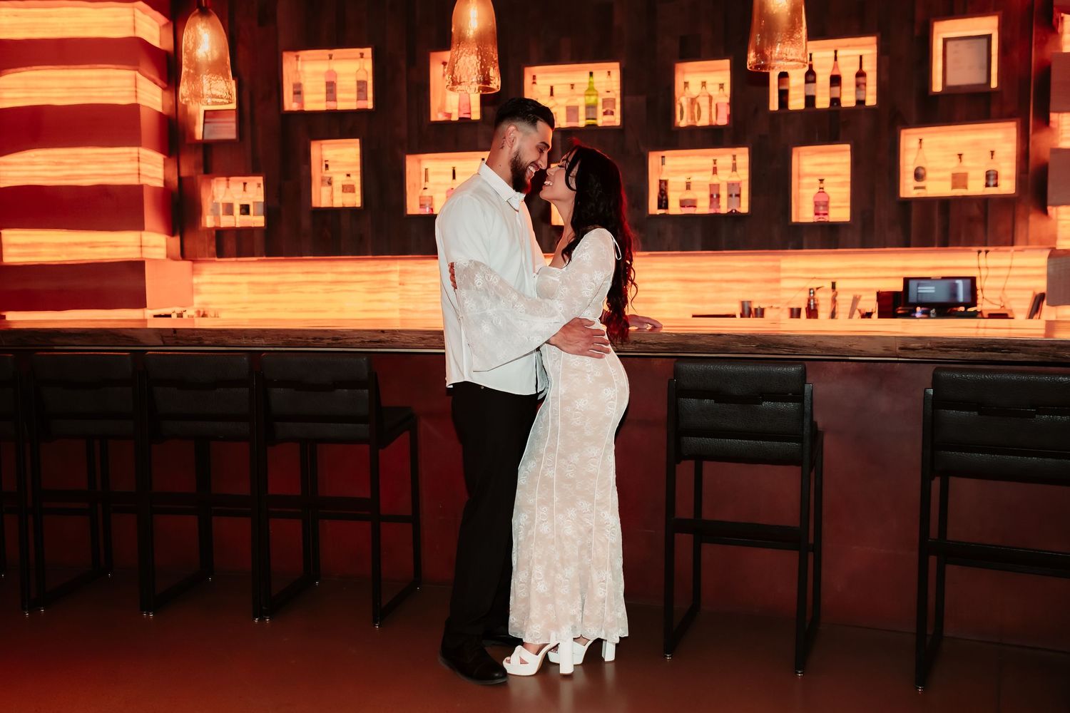 Couple embraces in front of illuminated bar with backlit bottle display.