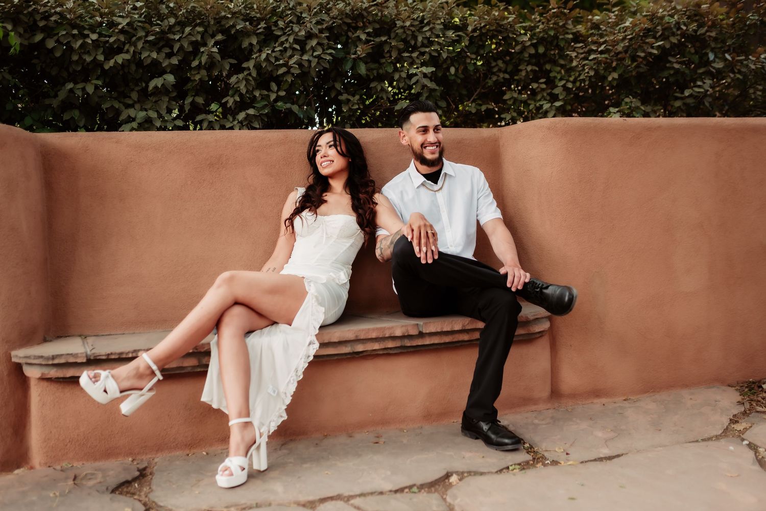 A couple in light colored outfits sits and poses together on a rustic adobe bench.