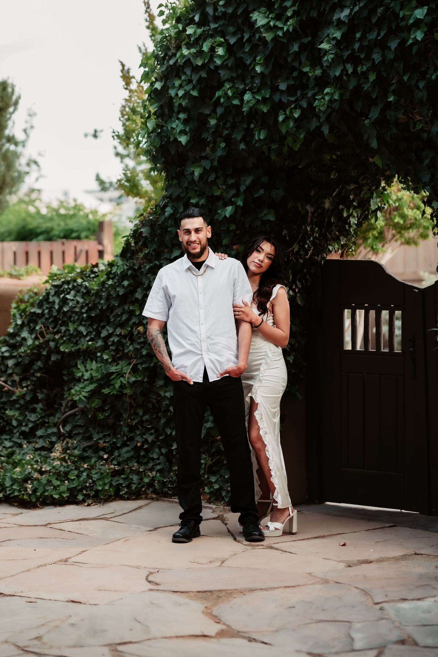 A stylish couple poses playfully on stone pavers with greenery and a wooden gate in the background.