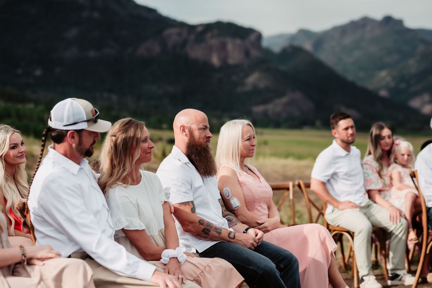Wedding guests seated in white chairs on grass with mountains in background.