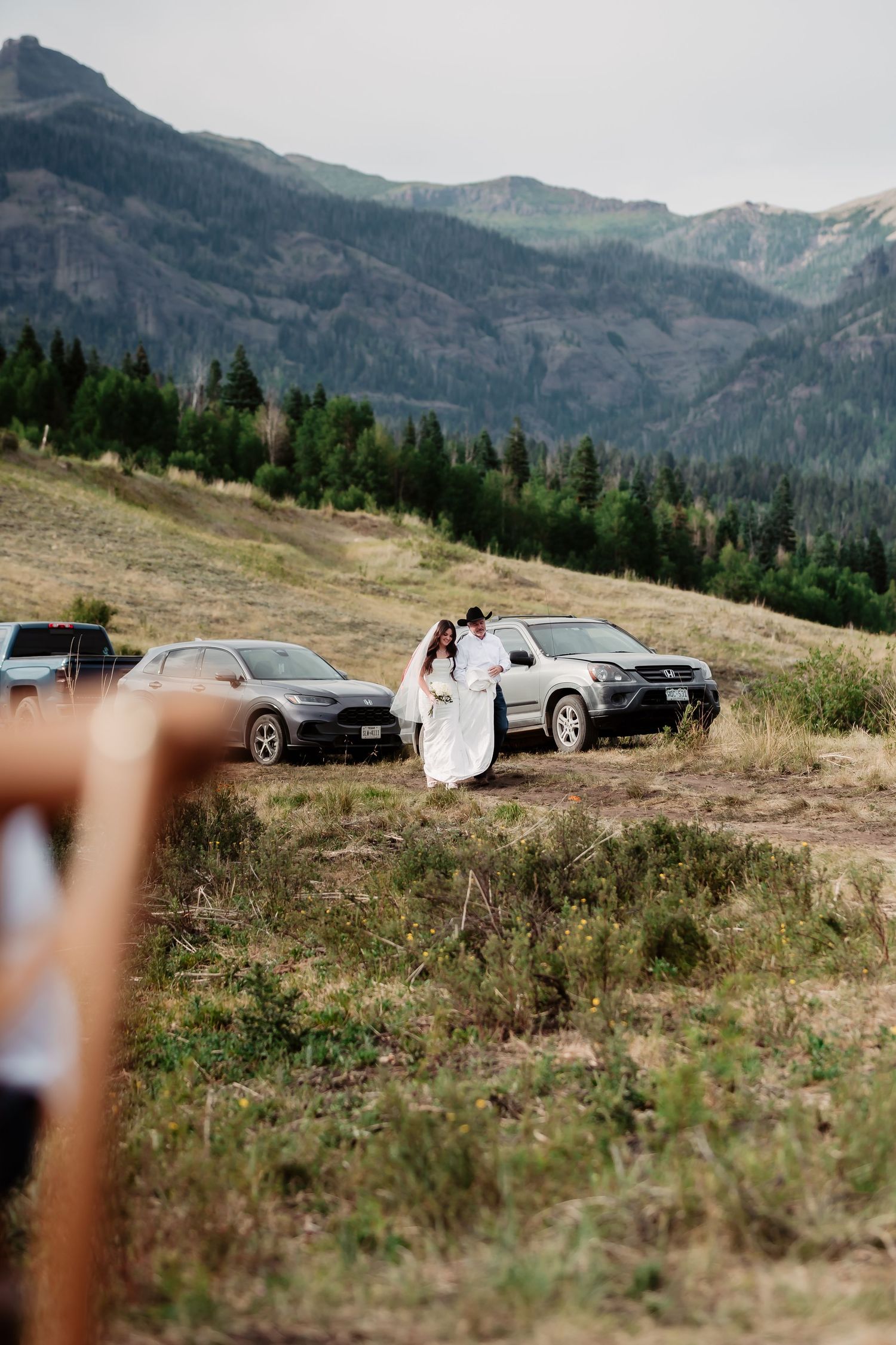 A couple poses in front of parked cars on a mountaintop with evergreen forest views.