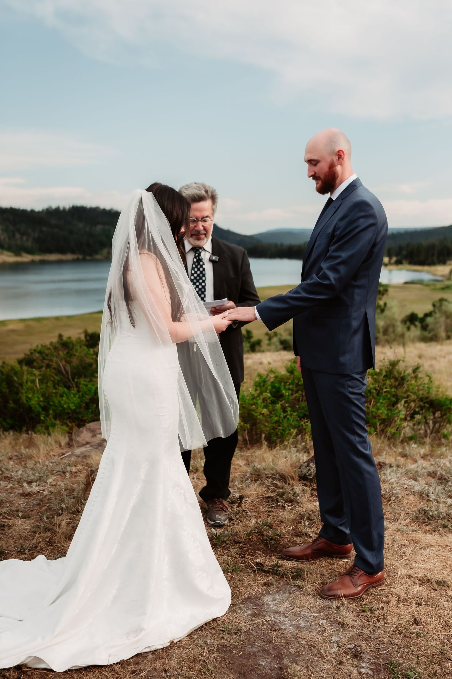 A wedding ceremony takes place on a scenic hillside overlooking mountains and water.