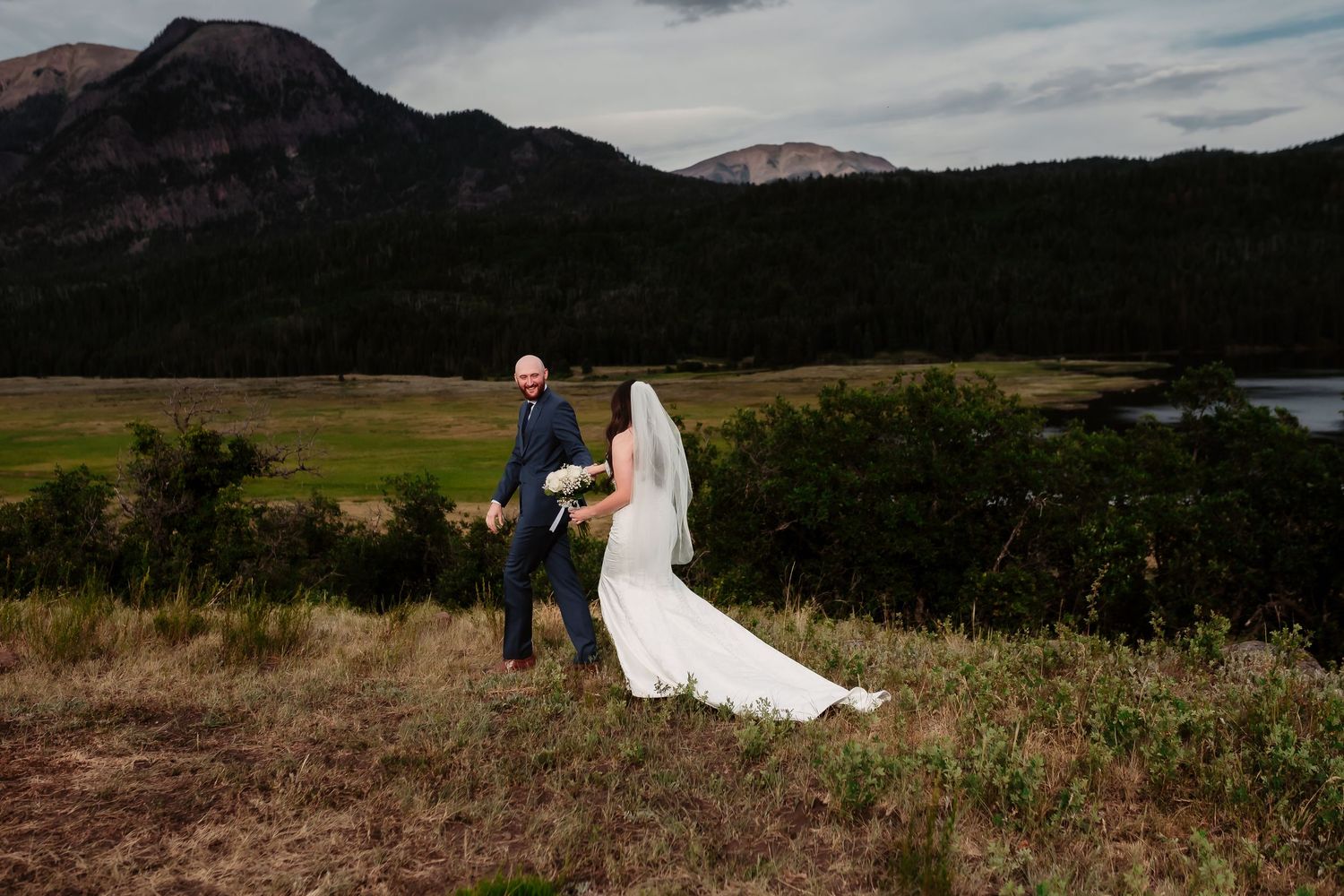 Bride in flowing white dress walks with companion against dramatic mountain and lake backdrop.