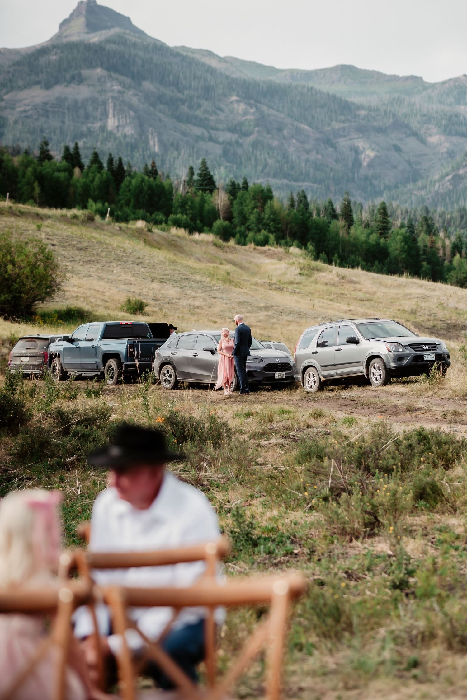Cars parked on a hillside with mountain peaks and evergreen forest in the background on a sunny day.