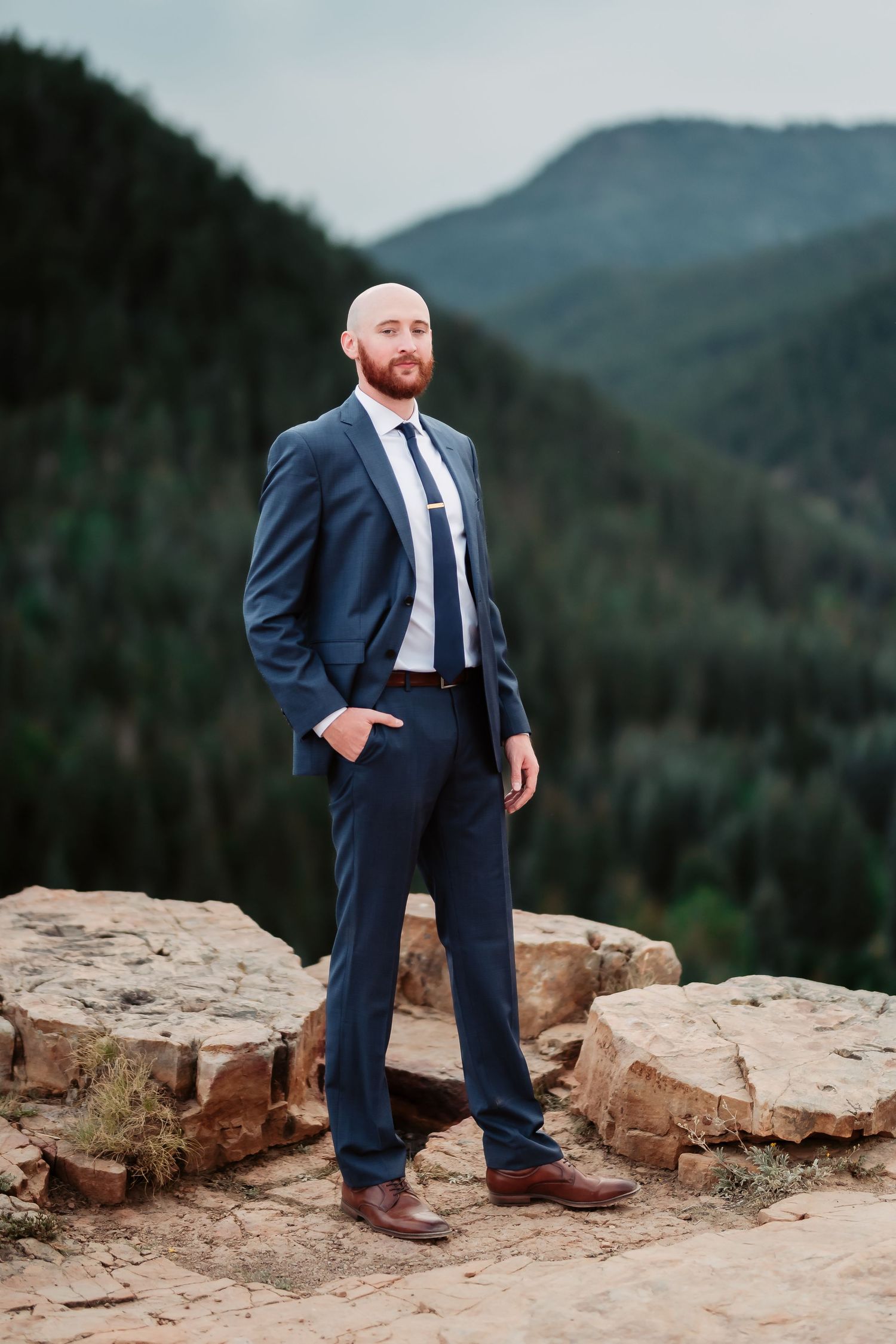 A groom in a navy suit stands confidently on rocky terrain with mountains in the background.