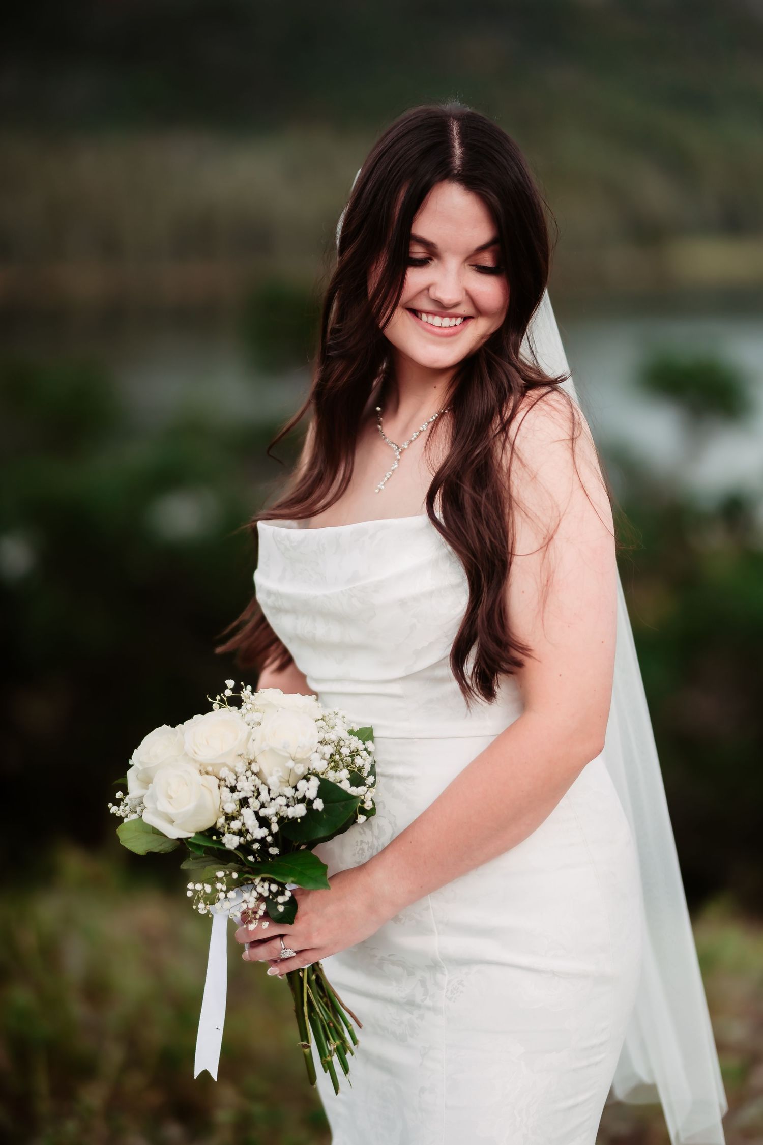 Bride in white dress holds bouquet of roses and baby's breath against mountain backdrop.