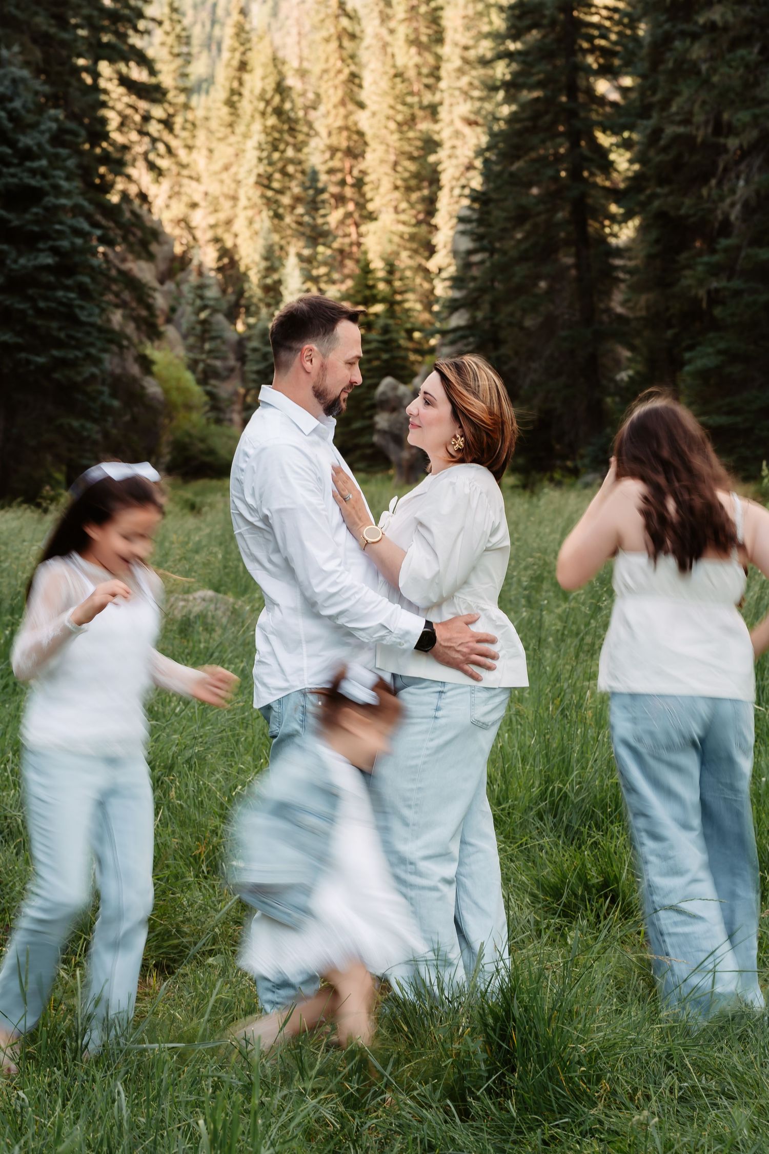 A family wearing coordinating white and light blue outfits dances together in a sunlit forest meadow.