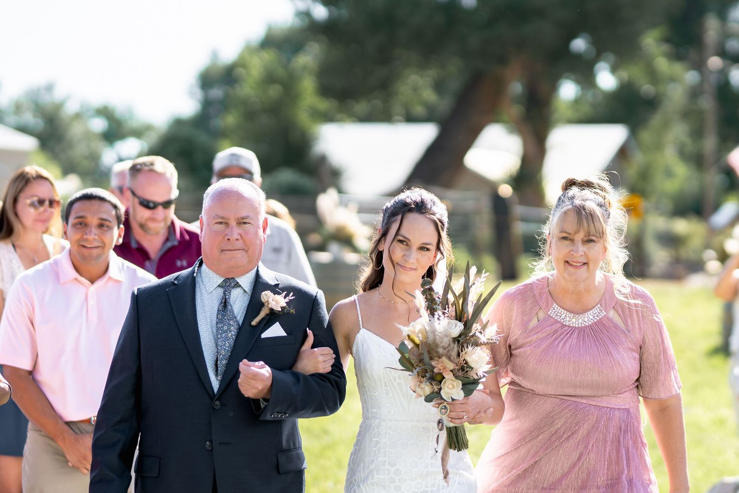 Wedding party walking down outdoor aisle during sunny ceremony with floral arrangements.