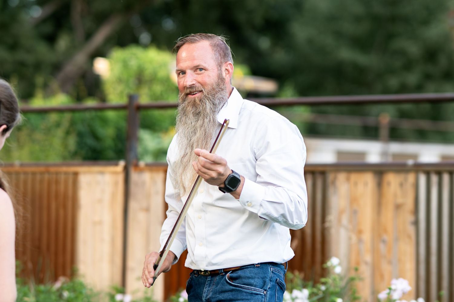 Man with long grey beard wearing white button-down shirt stands outdoors near wooden fence.