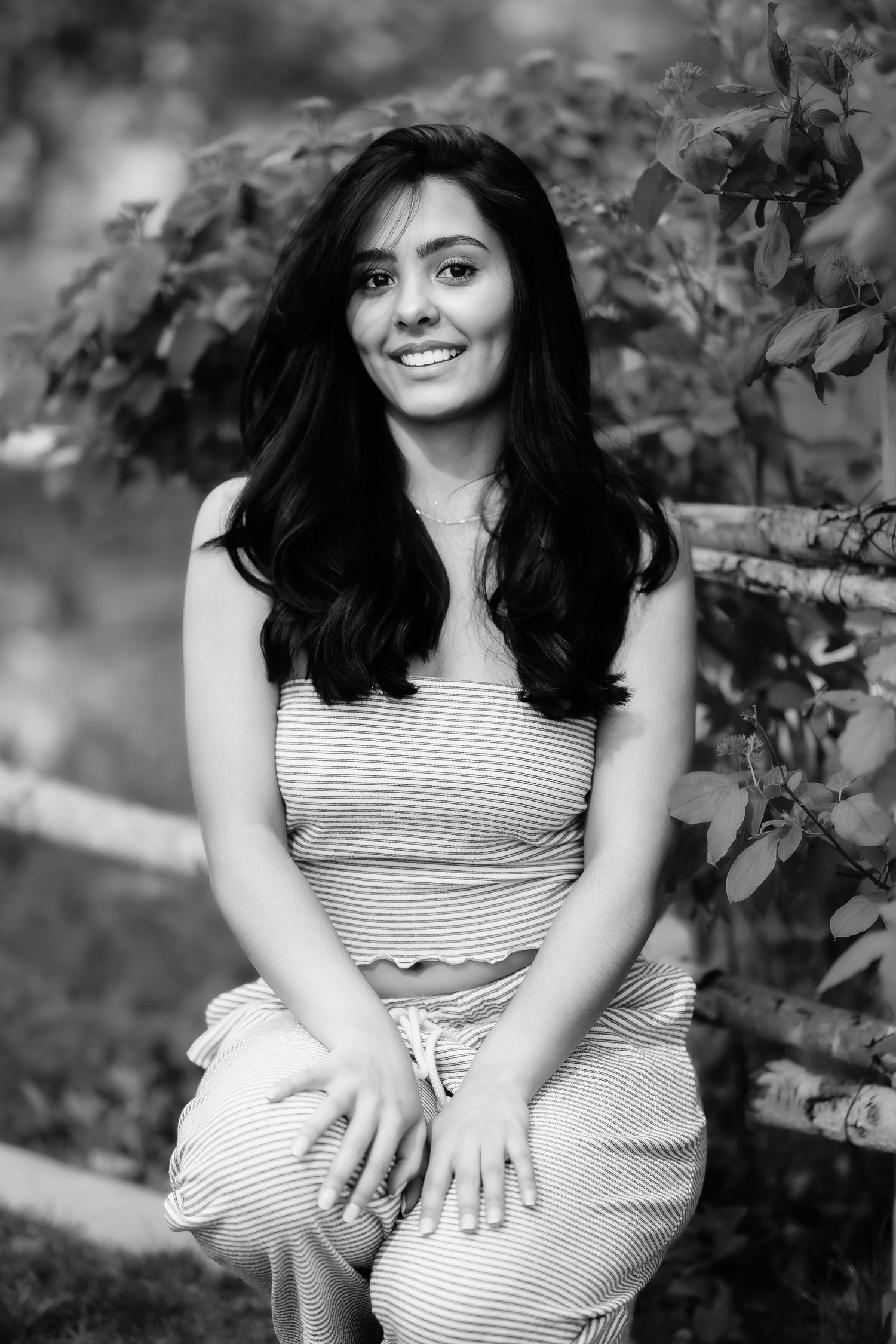 Monochrome portrait of woman in striped outfit sitting and smiling surrounded by garden leaves.