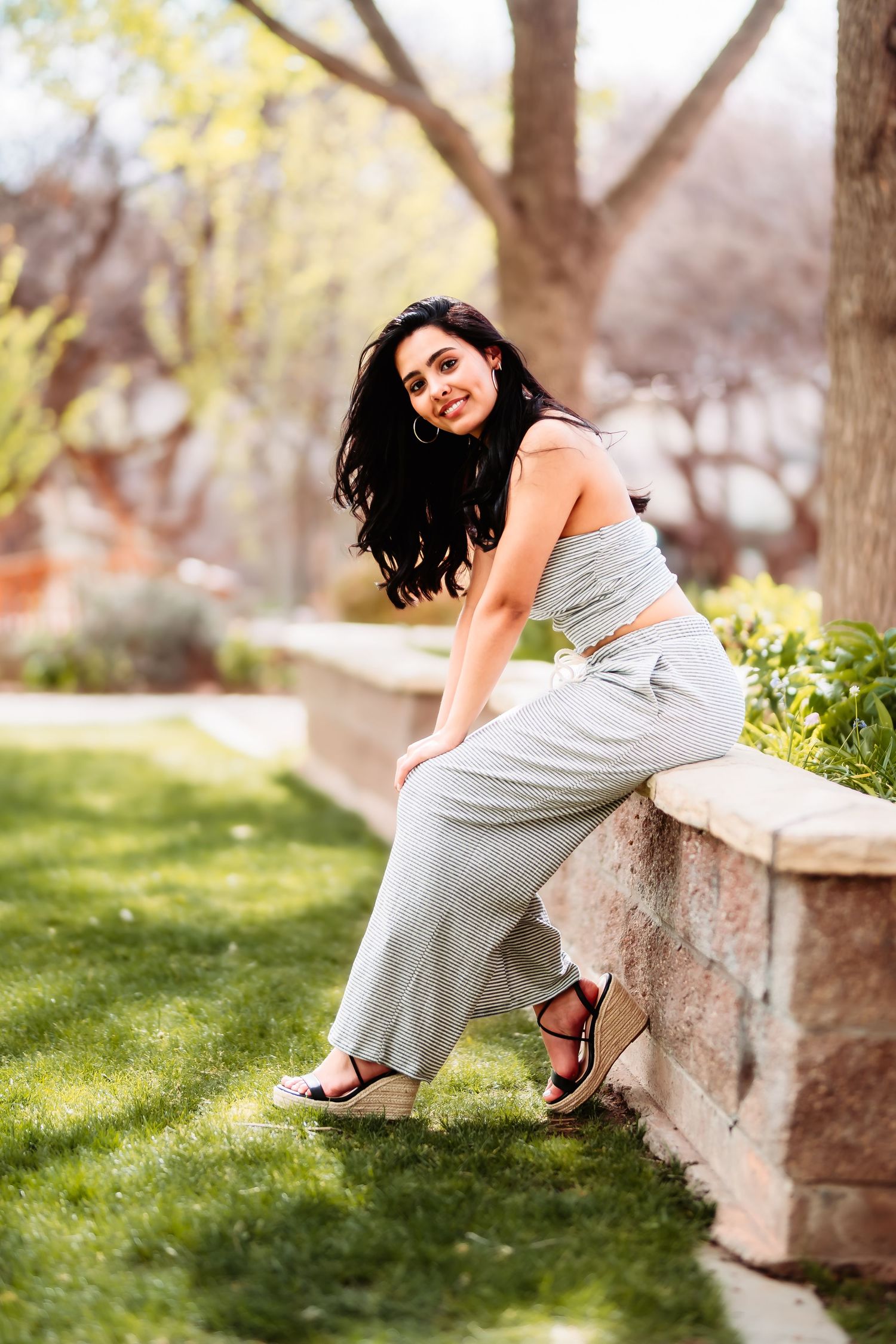 A casual portrait of someone in a grey dress sitting on a stone wall in a garden setting.