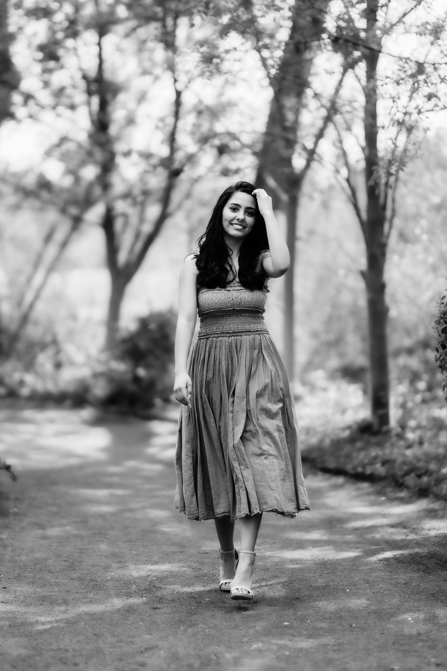 Black and white portrait of a person in a midi dress walking along a tree-lined path in a dreamy forest setting.