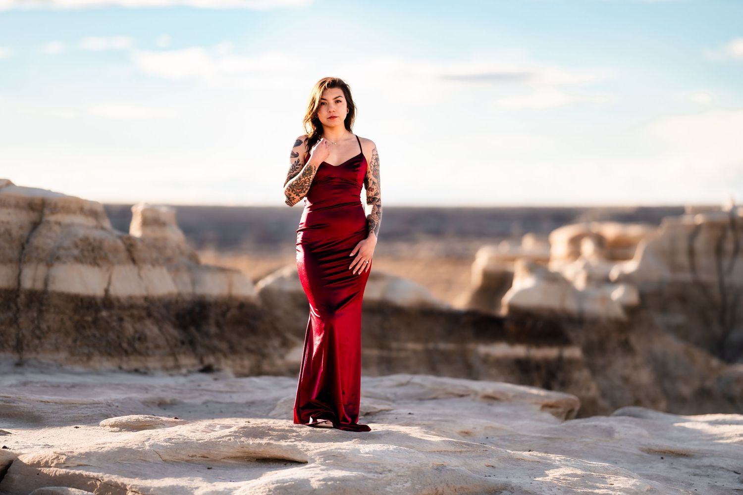 A woman in a flowing red dress stands elegantly on rocky terrain against a desert backdrop.