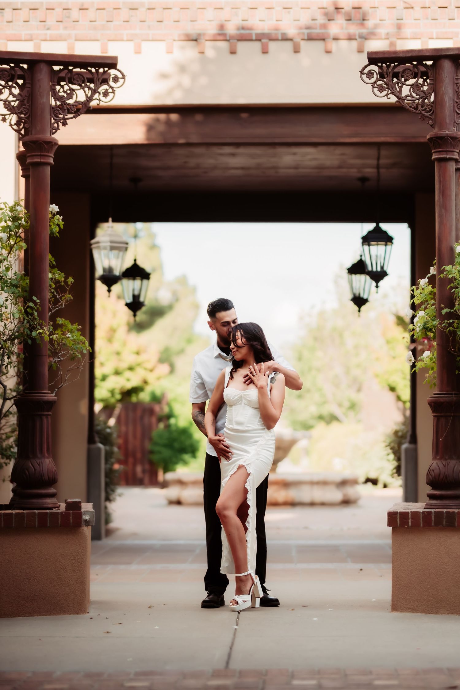 Romantic couple poses underneath a covered outdoor pergola with hanging lanterns at sunset.