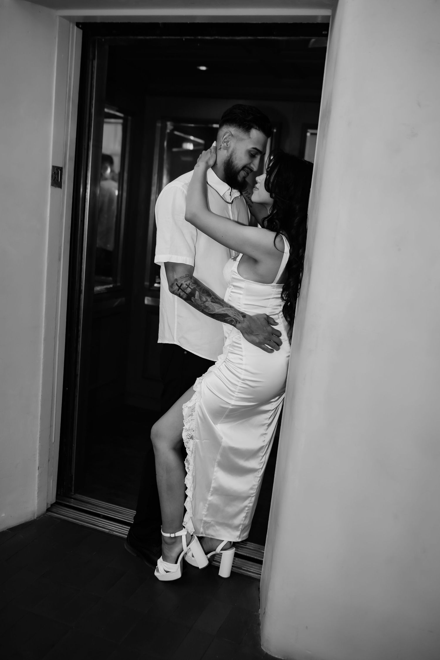 Black and white photo of couple in intimate embrace in doorway, both wearing white outfits.