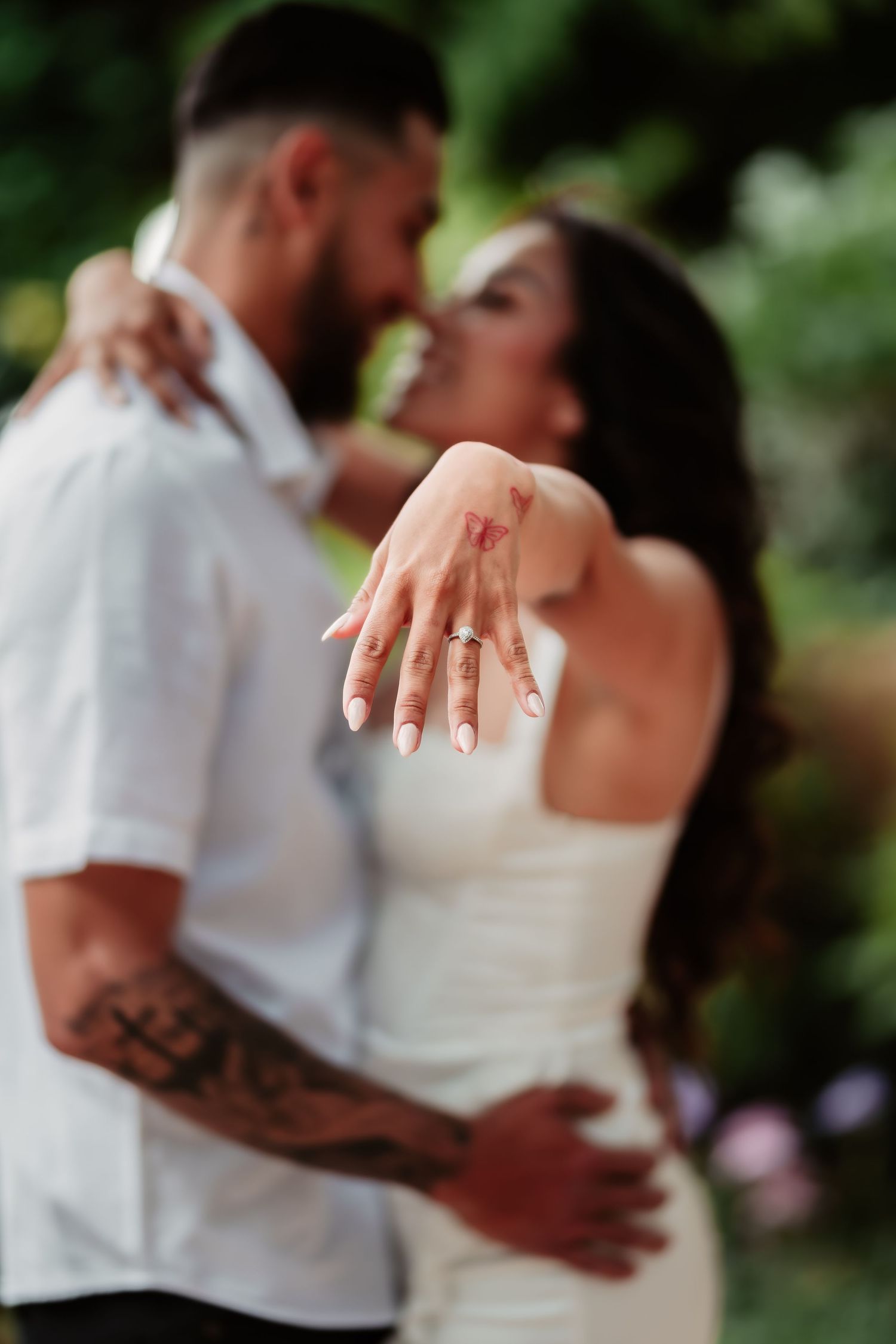 Newly engaged couple showing off engagement ring in romantic embrace against green foliage background.