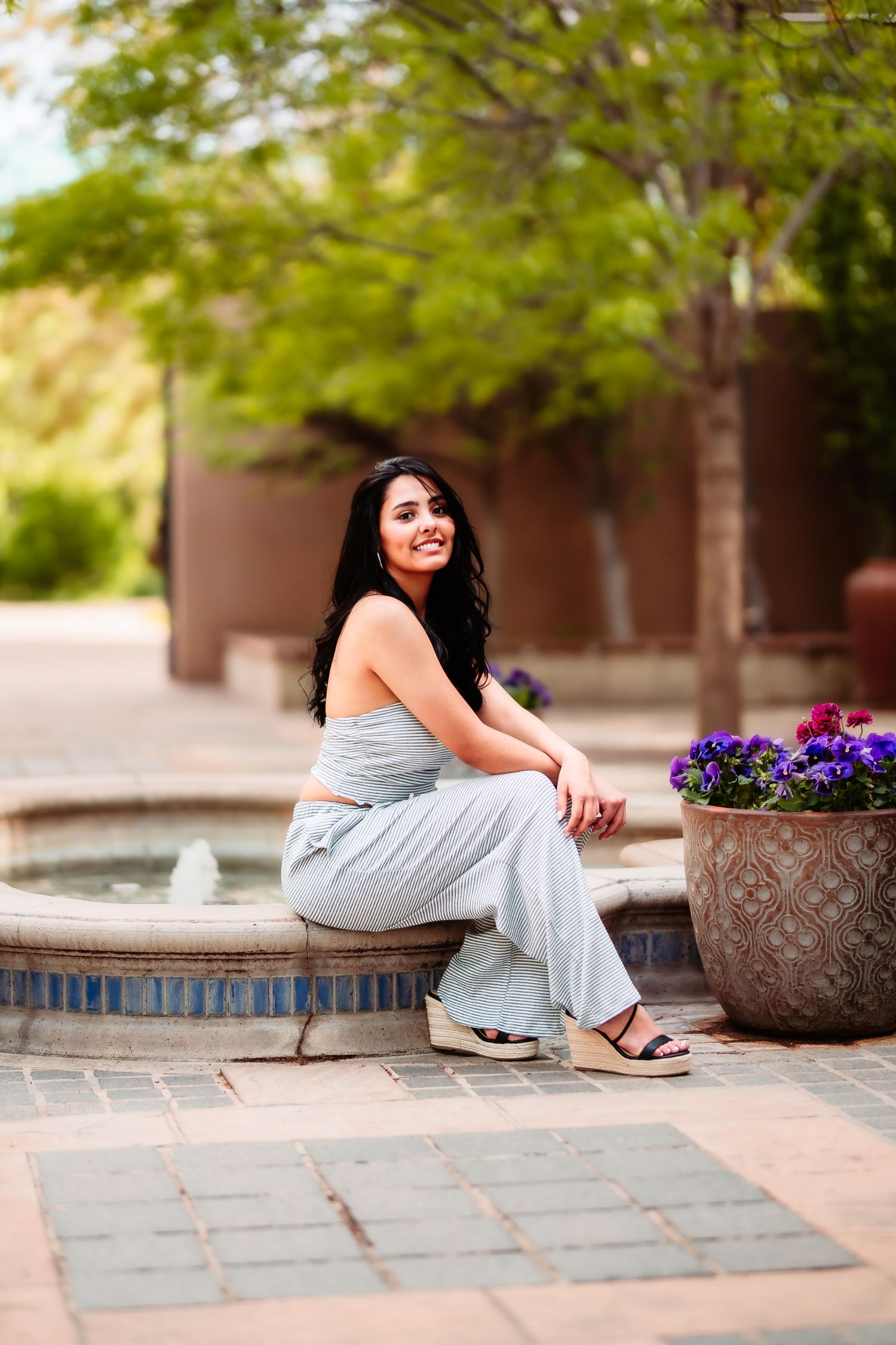 Woman in casual attire sitting by fountain with purple flowers and architectural elements in background.