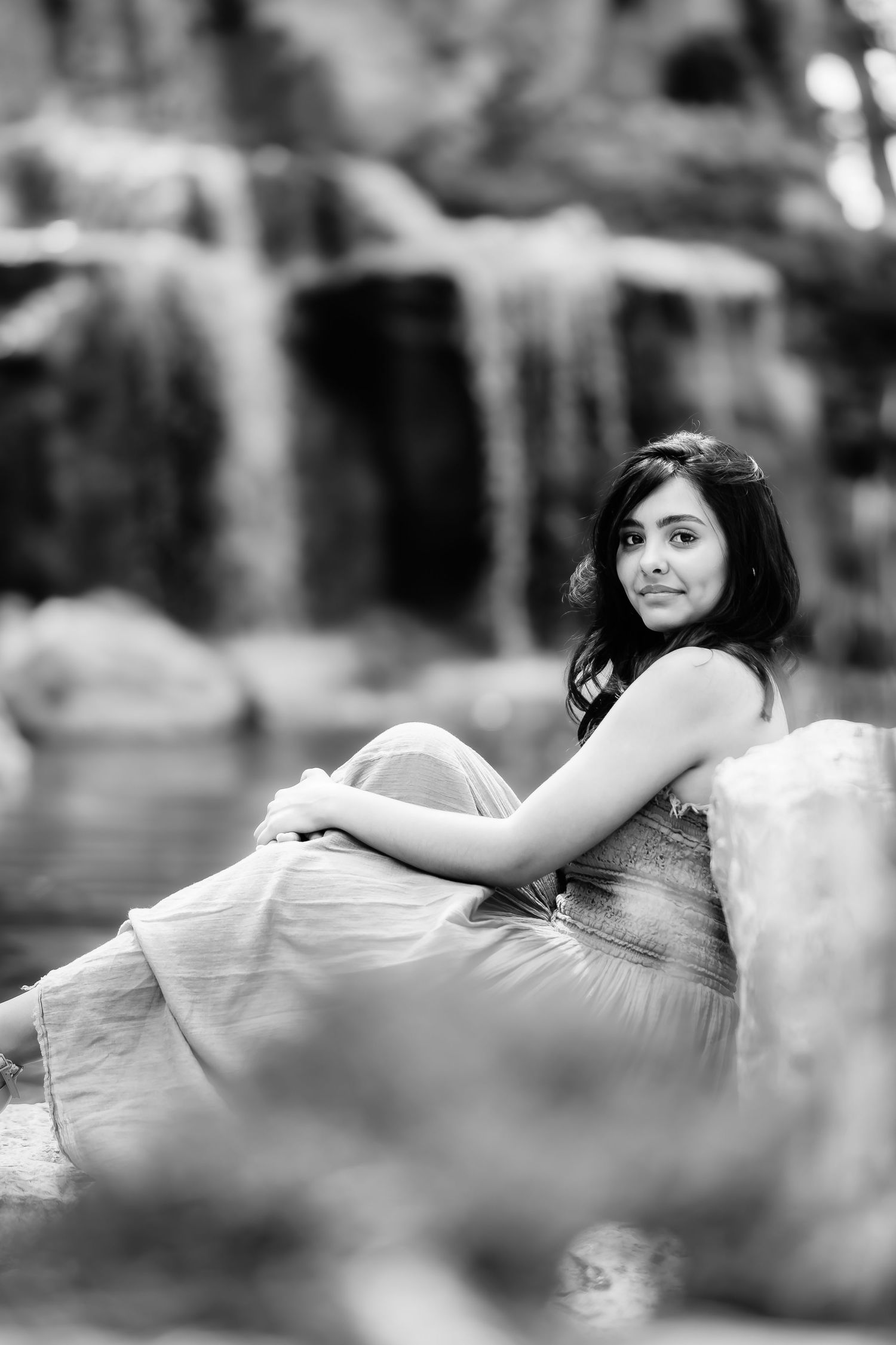 A black and white portrait by a waterfall captures someone in a light dress resting on rocks.