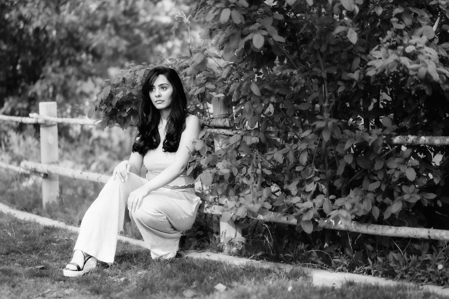 Artistic black and white portrait of woman crouching by rustic fence.
