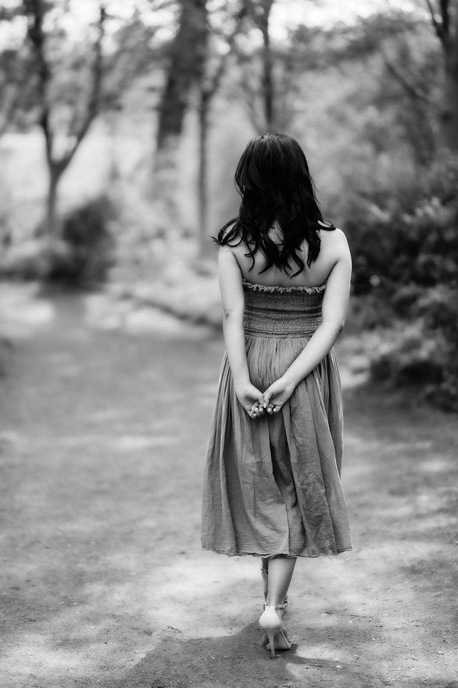 A black and white photo of someone in a dress walking down a wooded path, shot from behind.