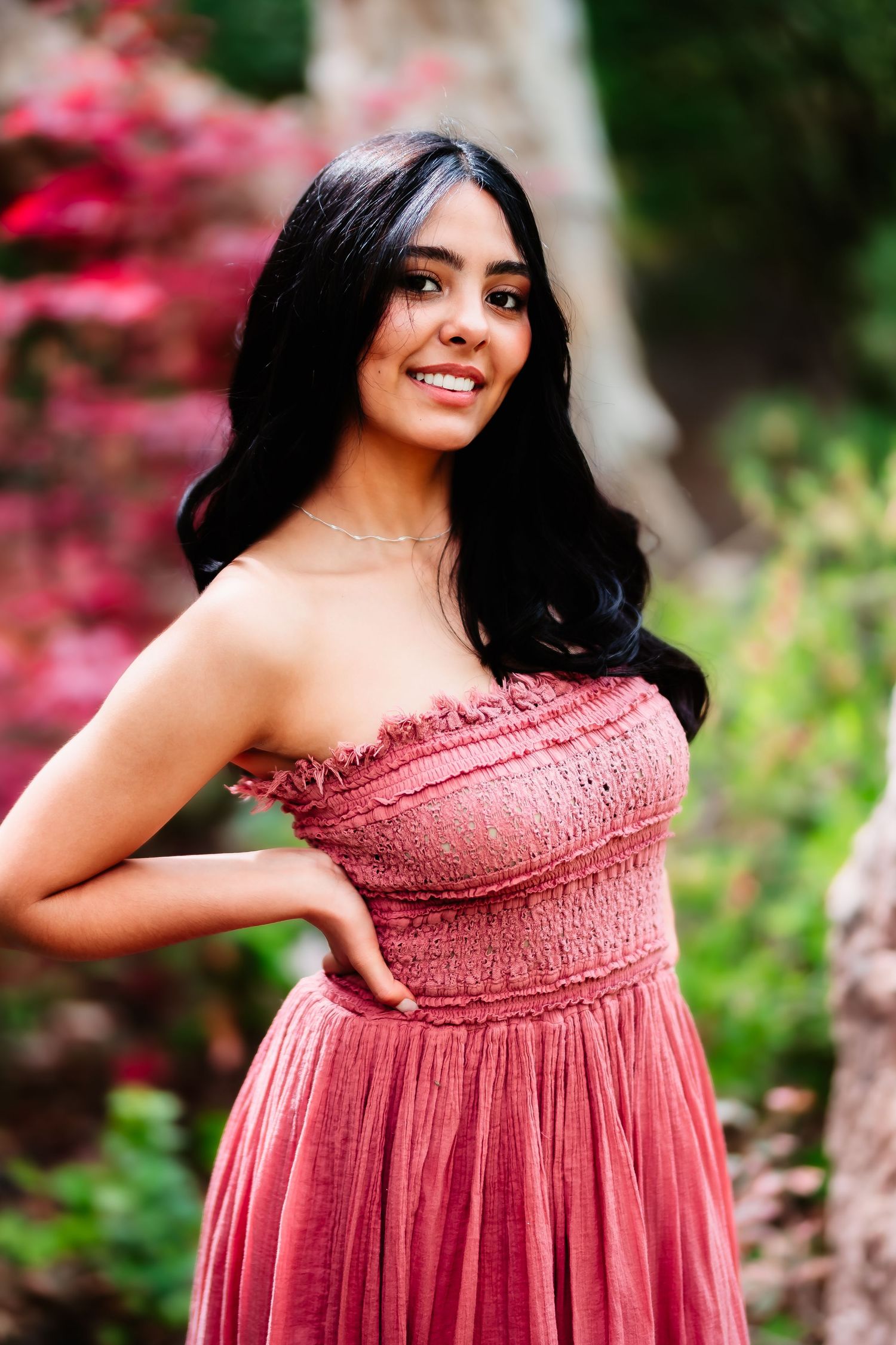 A woman in a pink strapless dress poses outdoors against vibrant foliage and flowers.
