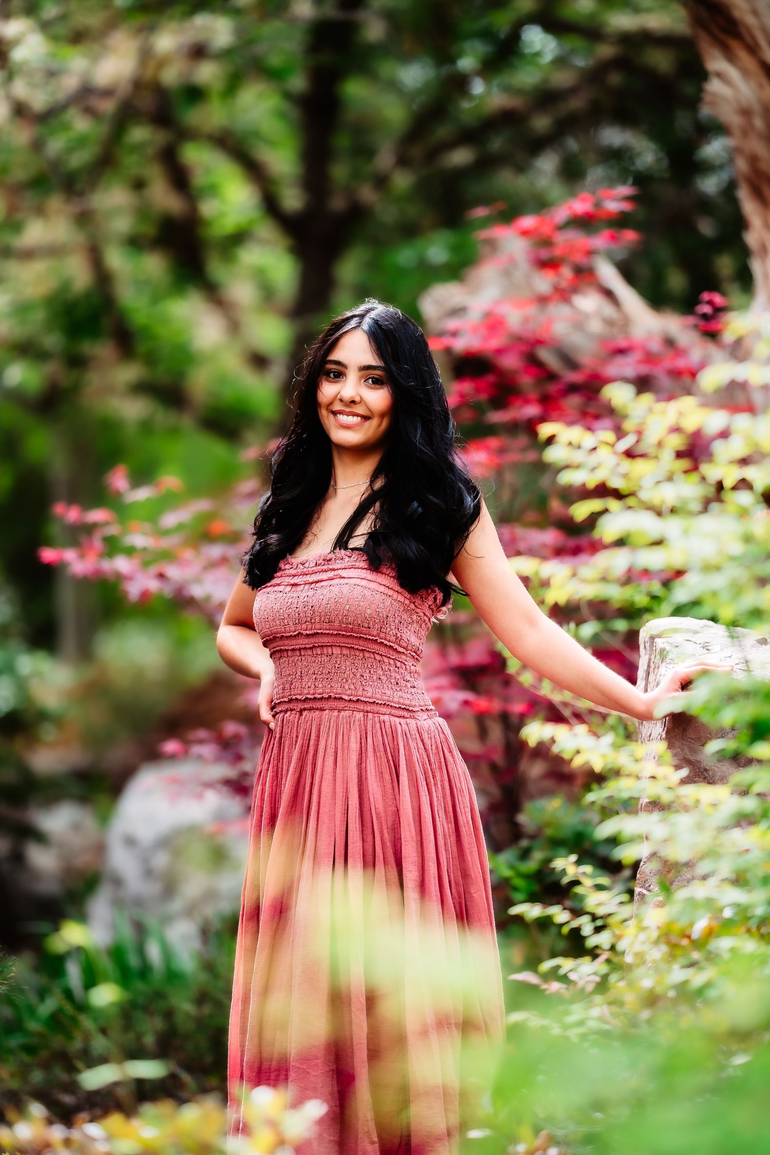 A pink ombre dress flows elegantly against vibrant red maple leaves and greenery in a garden setting.