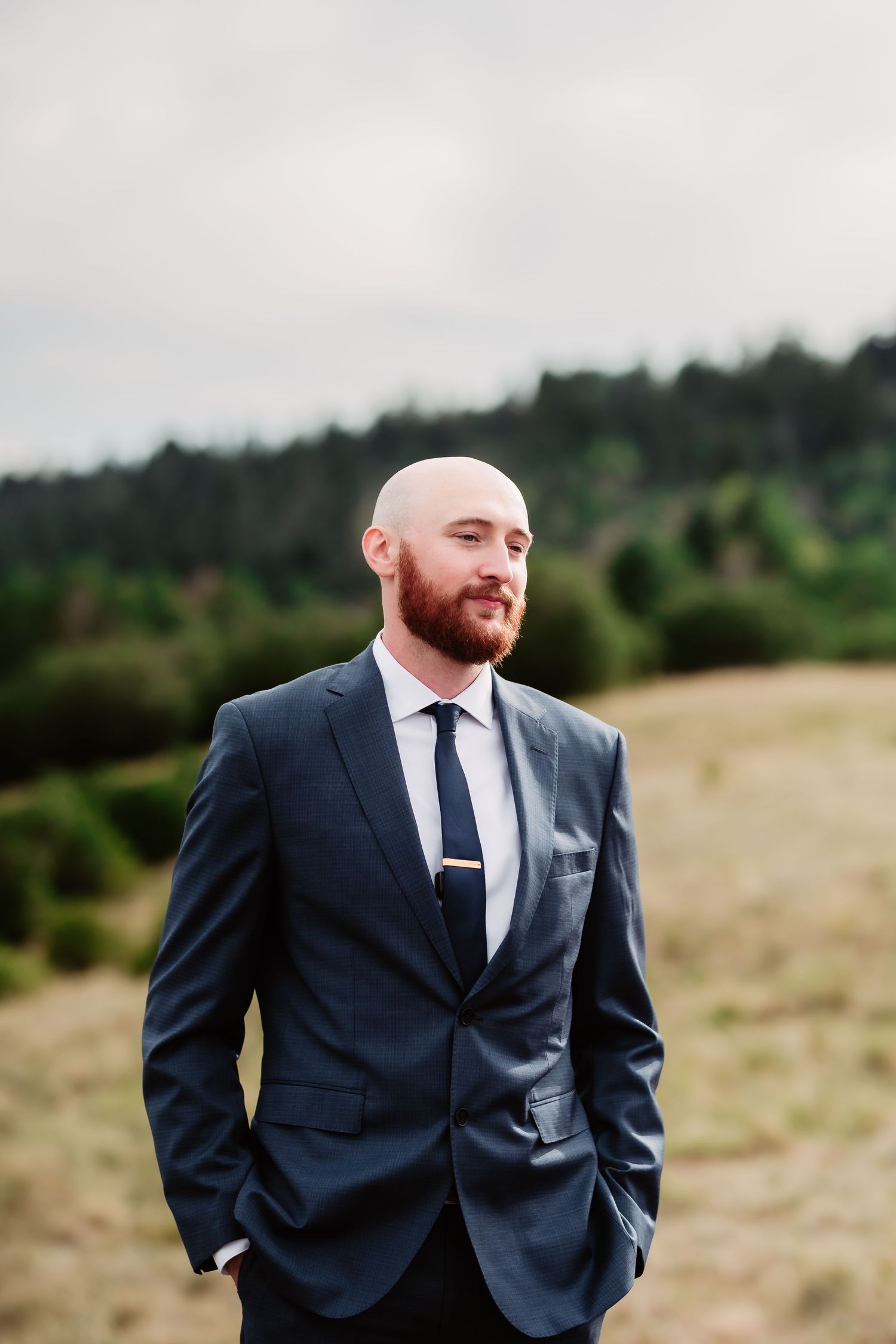A groom in a navy suit and tie stands outdoors against a natural backdrop with hills and forest.