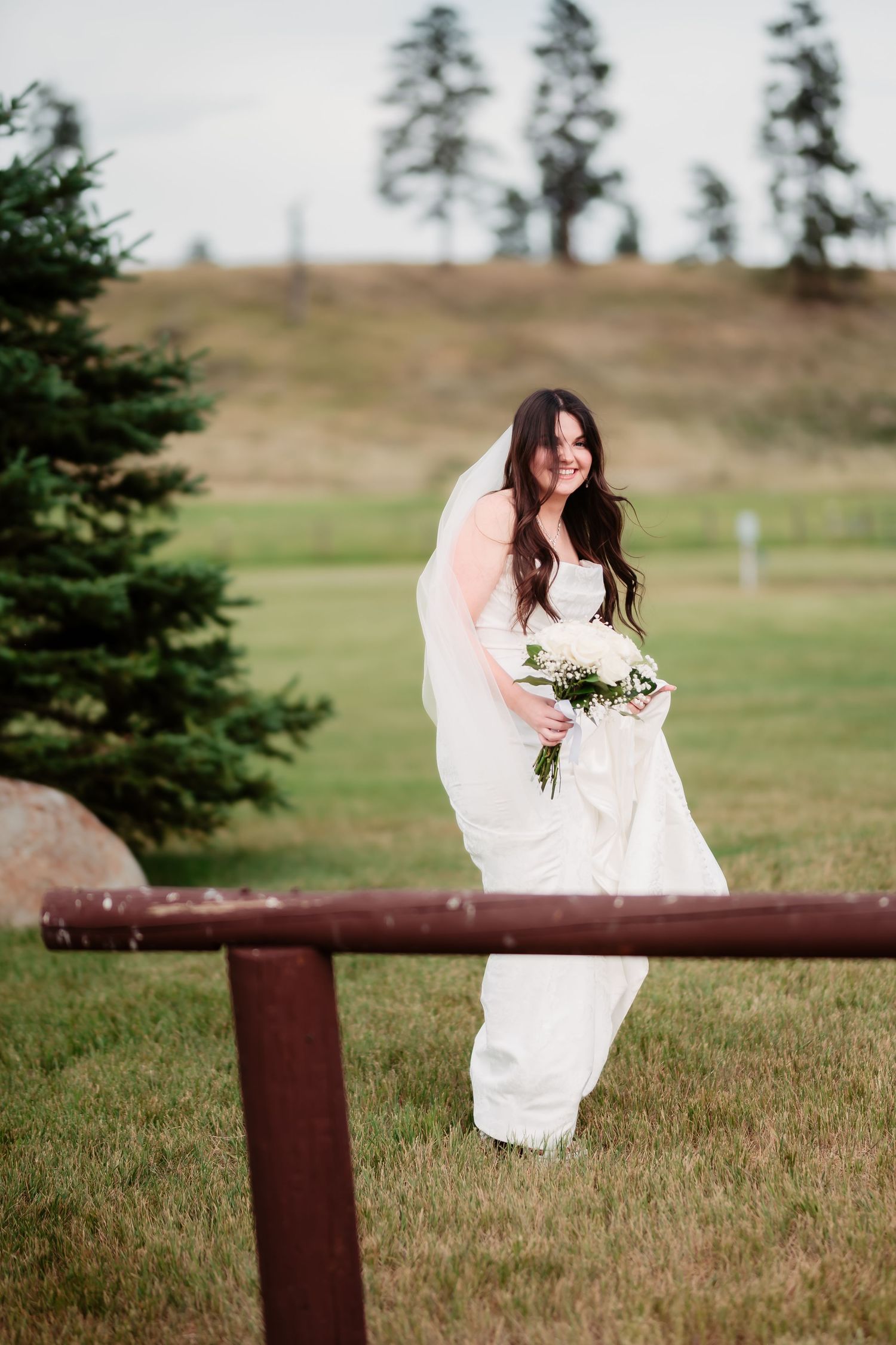 A bride in white dress poses by a rustic fence with mountains and evergreens in the background.