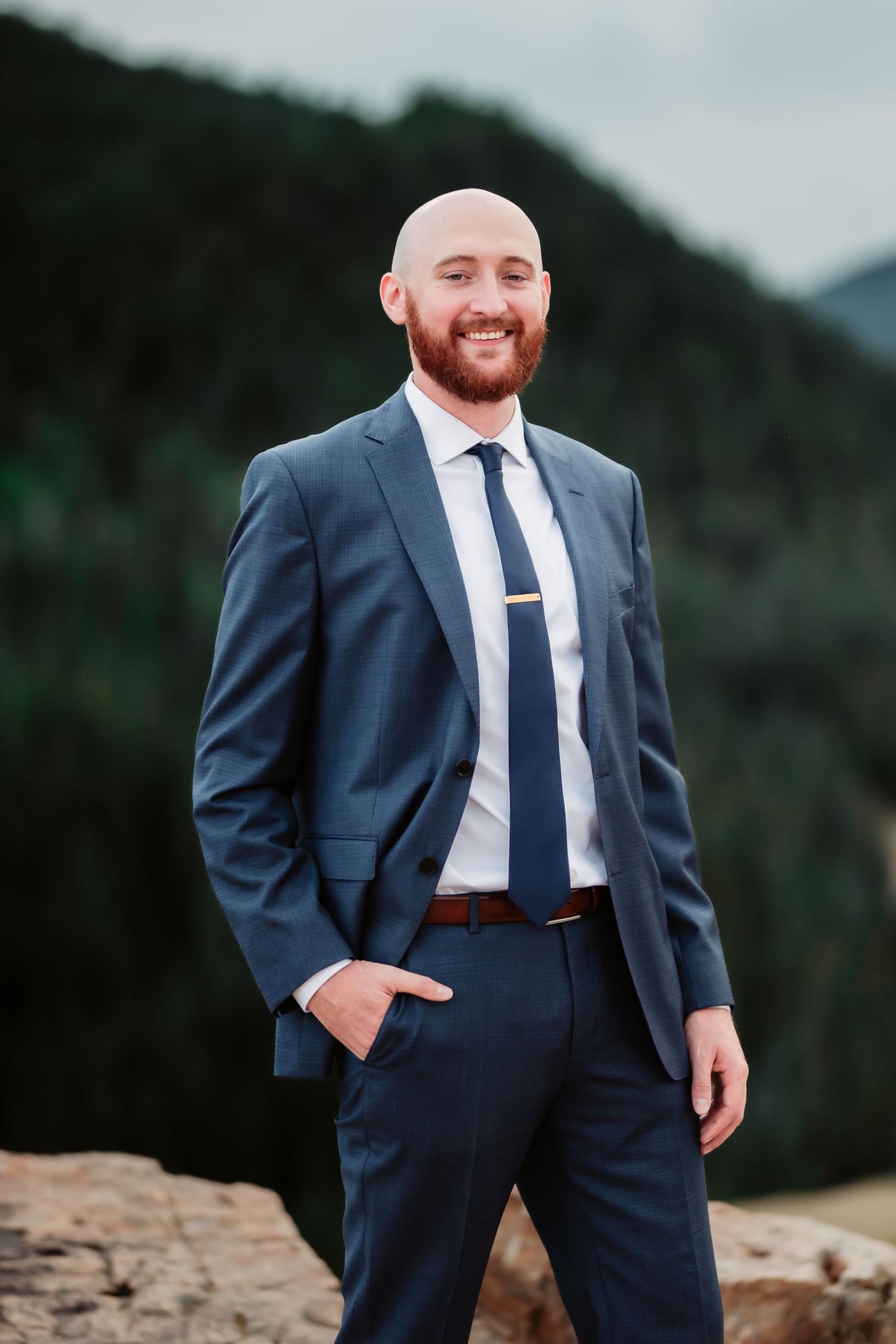 A groom in a tailored navy suit poses on a mountain overlook during his wedding day.