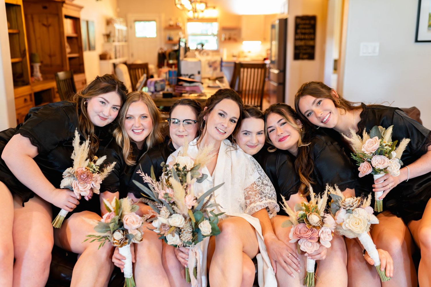 Wedding party wearing matching black robes poses while seated together holding floral bouquets in warm-lit room.