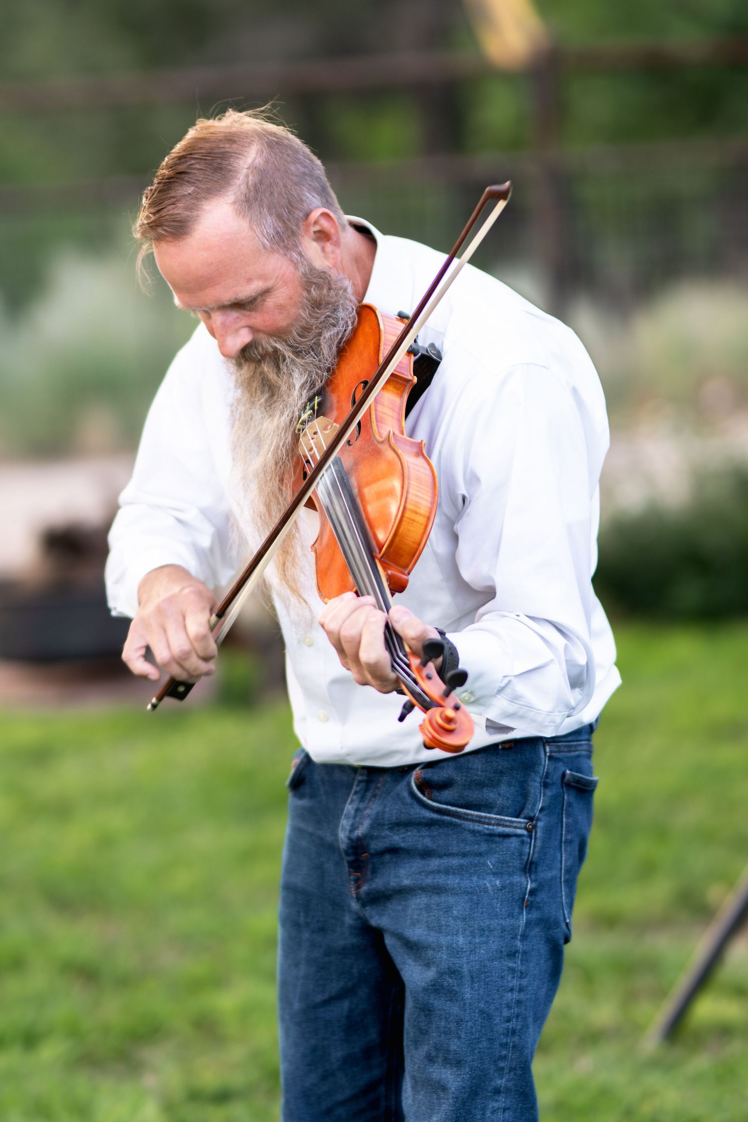 A musician plays violin outdoors wearing a white shirt and jeans with a long beard in a garden setting.