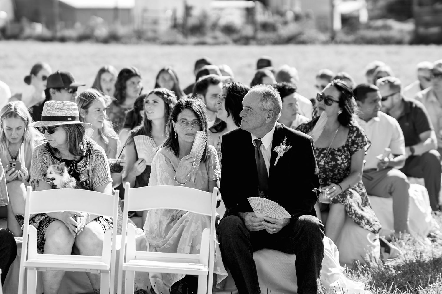 Black and white candid of wedding guests seated in rows of white chairs at outdoor ceremony.