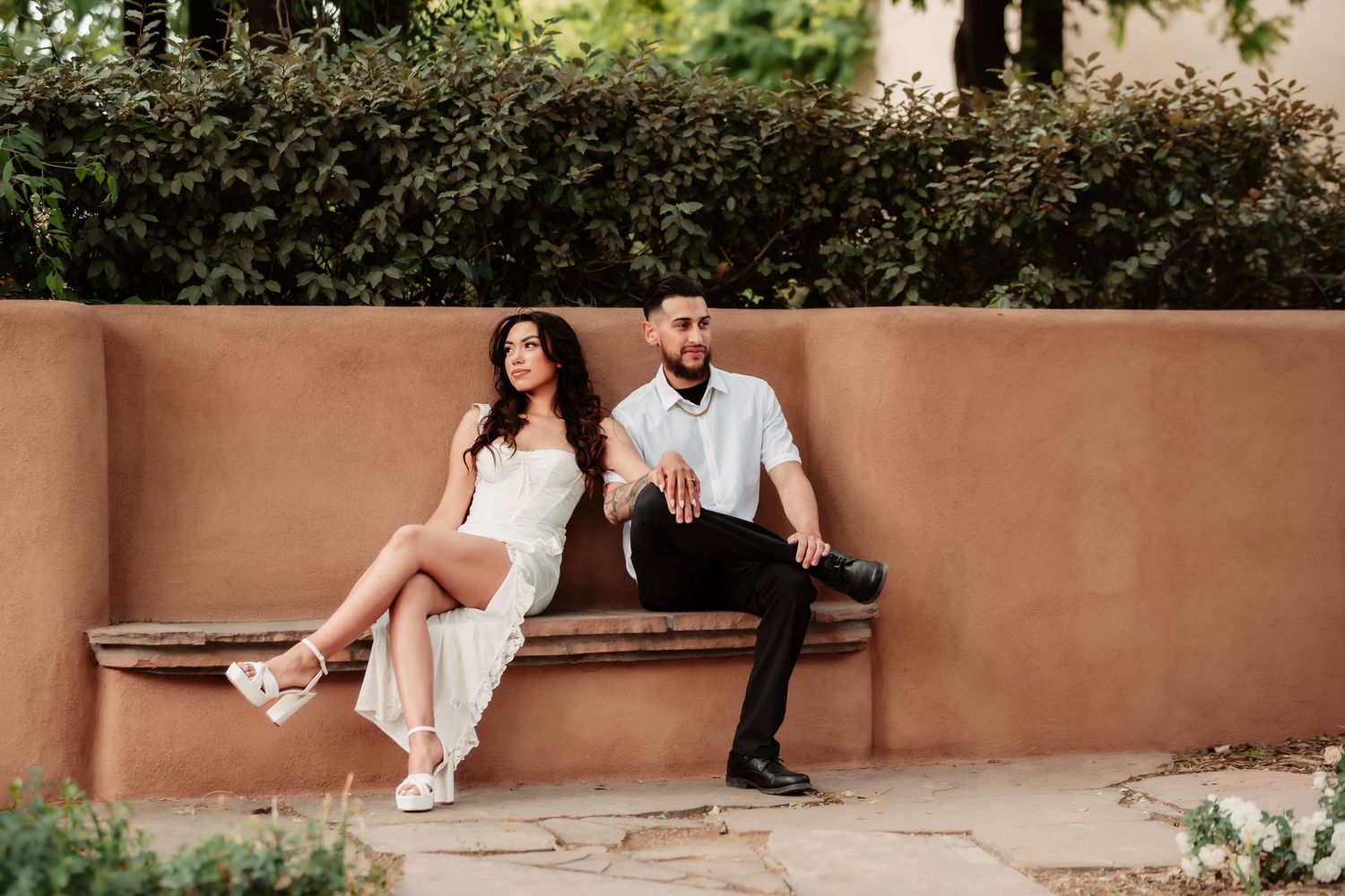 A stylish couple sits together on a terracotta colored bench in an outdoor garden setting.