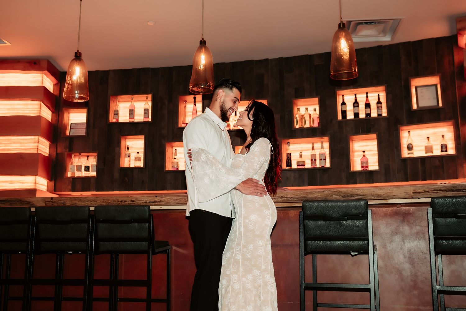 Couple embraces in front of illuminated bar shelving during romantic photoshoot.