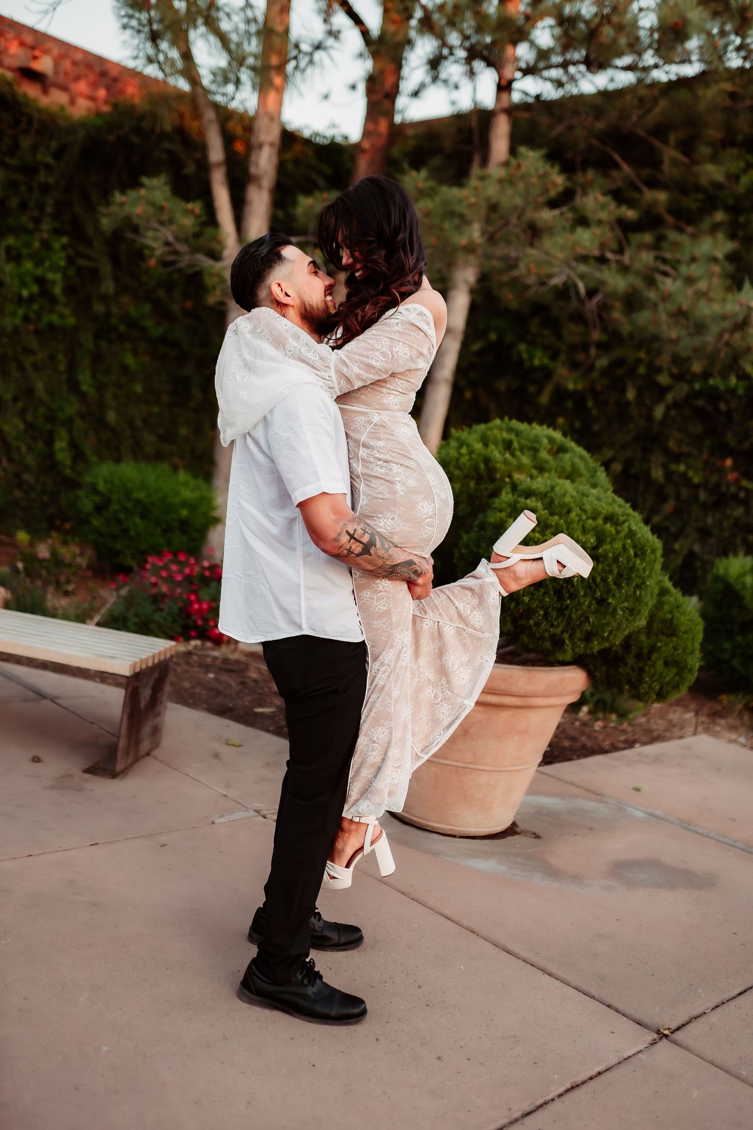 A couple shares a romantic moment in a garden setting at sunset, with the woman wearing a lace dress and lifting her leg.