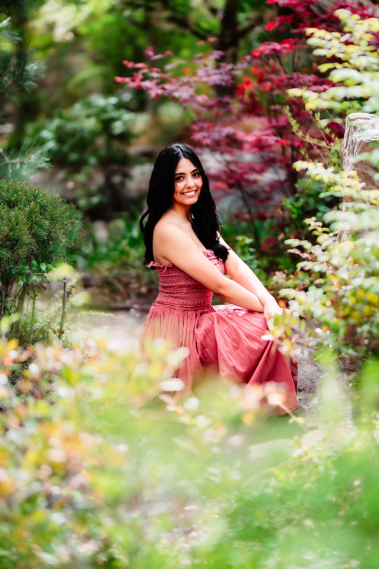 A woman in a rose-colored dress sits among colorful garden flowers and foliage.