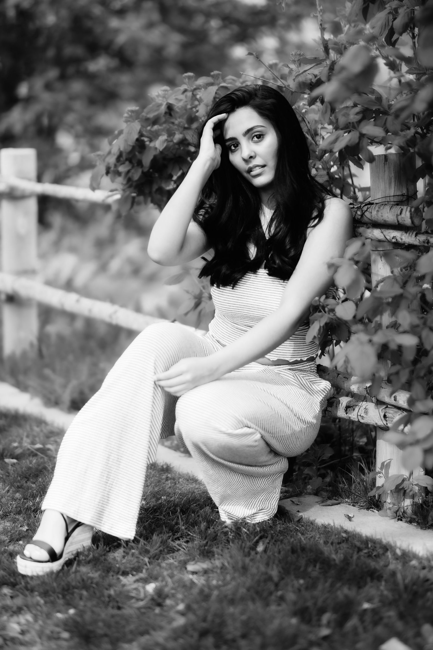 Woman in white outfit poses casually near wooden fence and foliage in monochrome portrait.