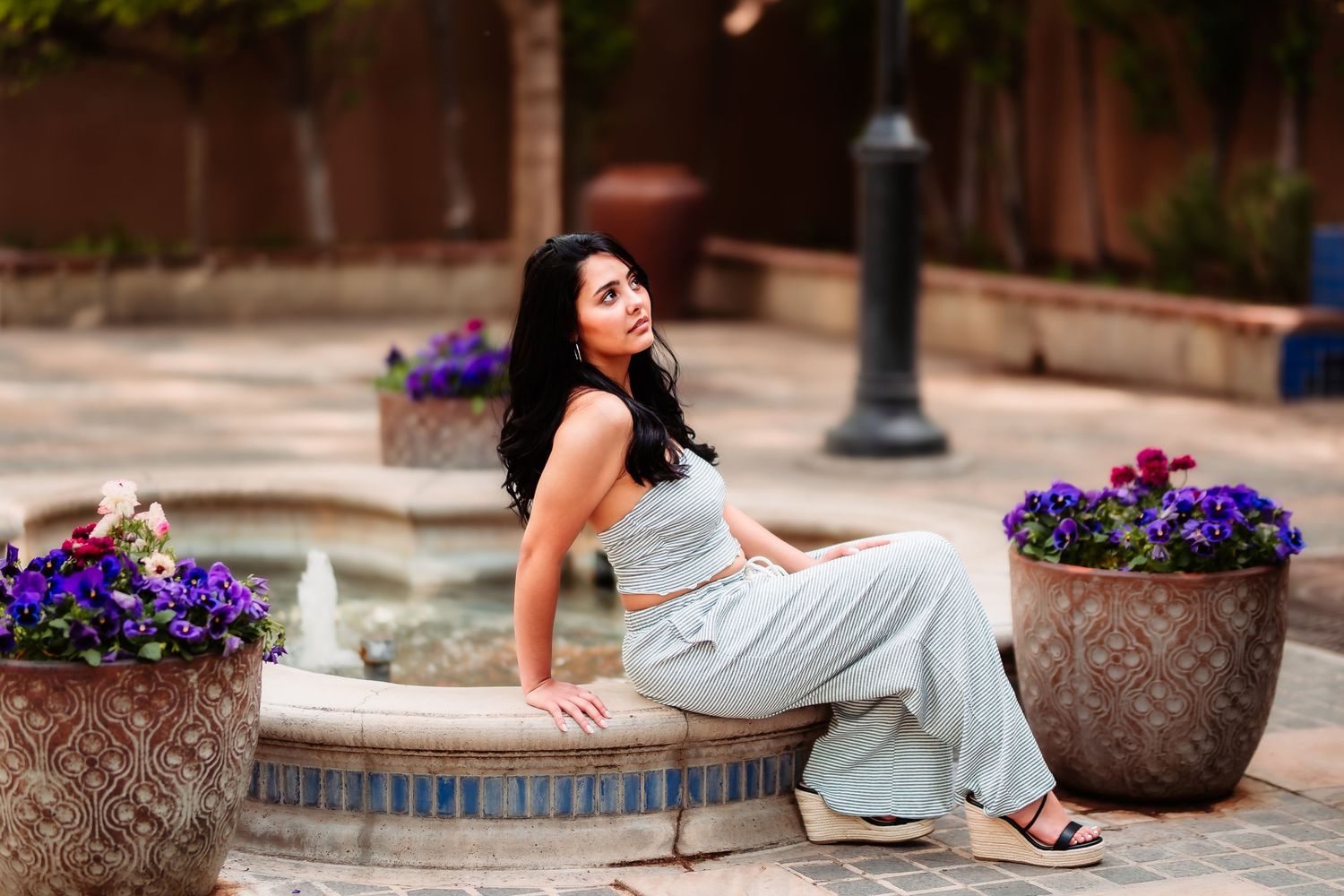 Woman in gray outfit sitting gracefully on stone ledge with purple flowers in decorative planters nearby.