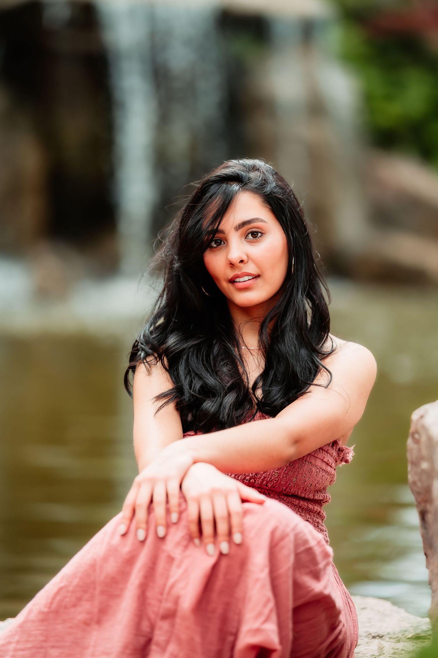 Woman in pink dress sitting casually by water feature with cascading waterfall in background.