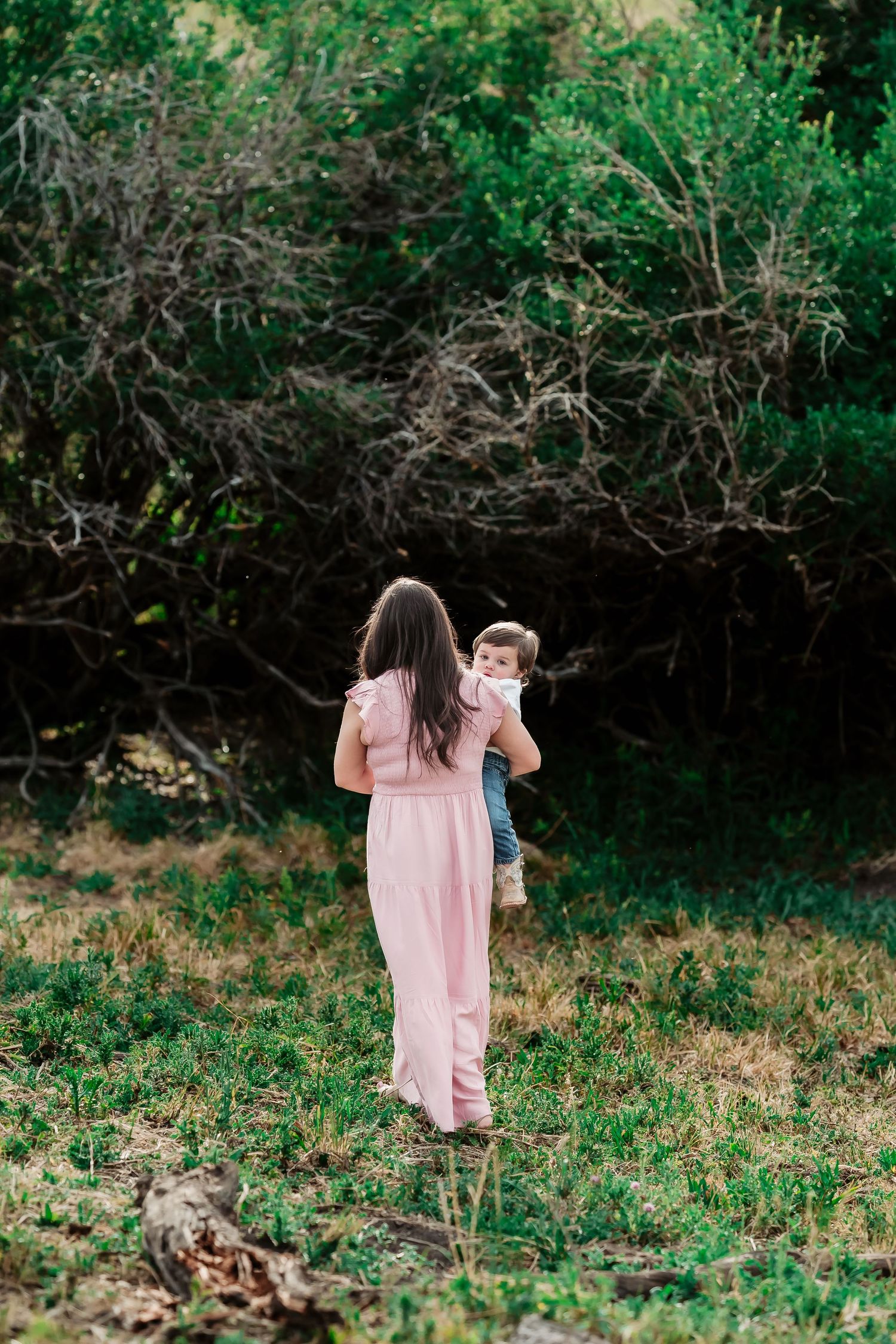 A woman in a pink dress walks through a grassy area carrying a small child.