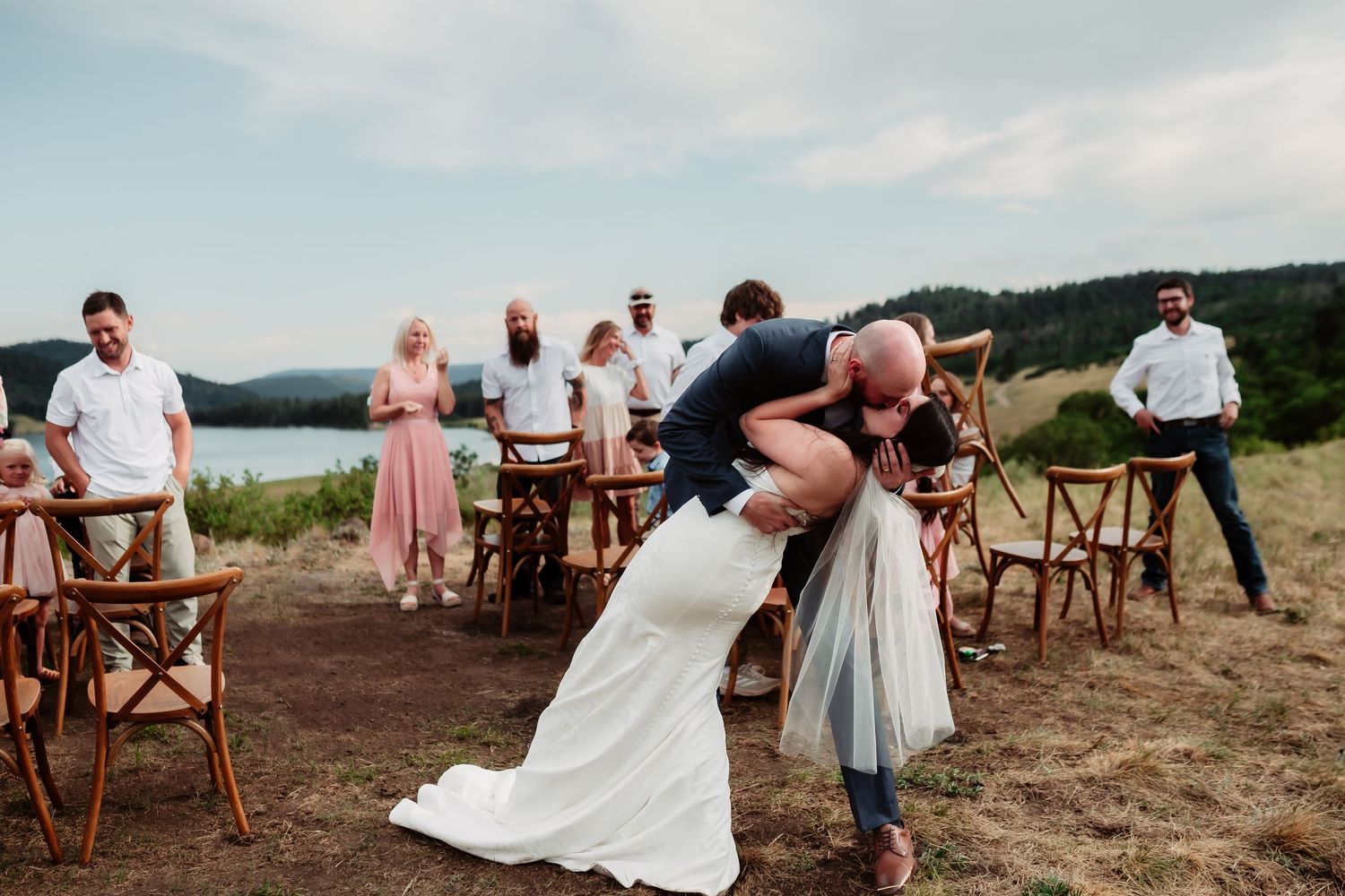Wedding party and guests gathered outdoors with wooden chairs set up on a rustic hilltop setting at sunset.