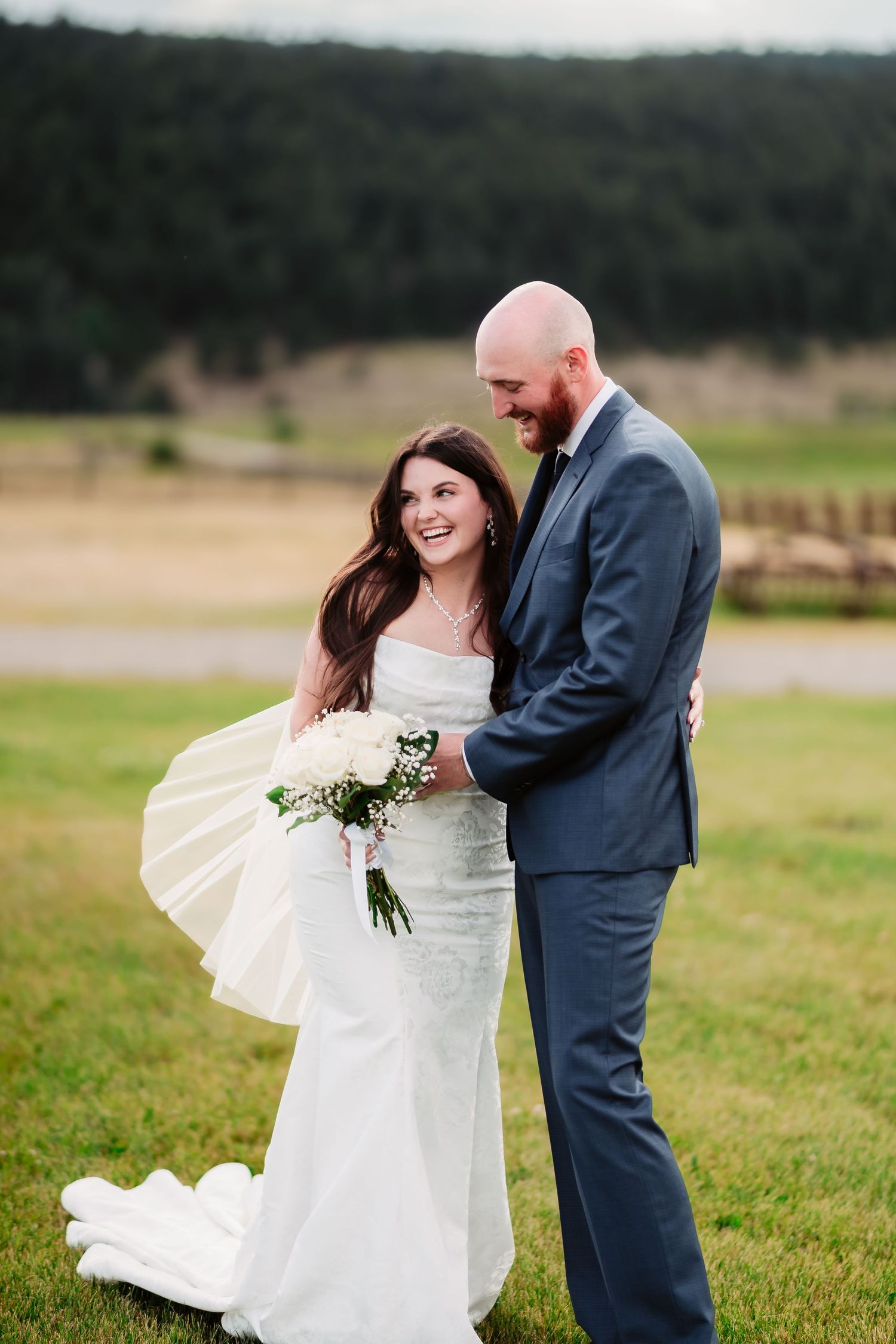 A couple shares a joyful moment on their wedding day, standing in a grassy field with mountains in the background.