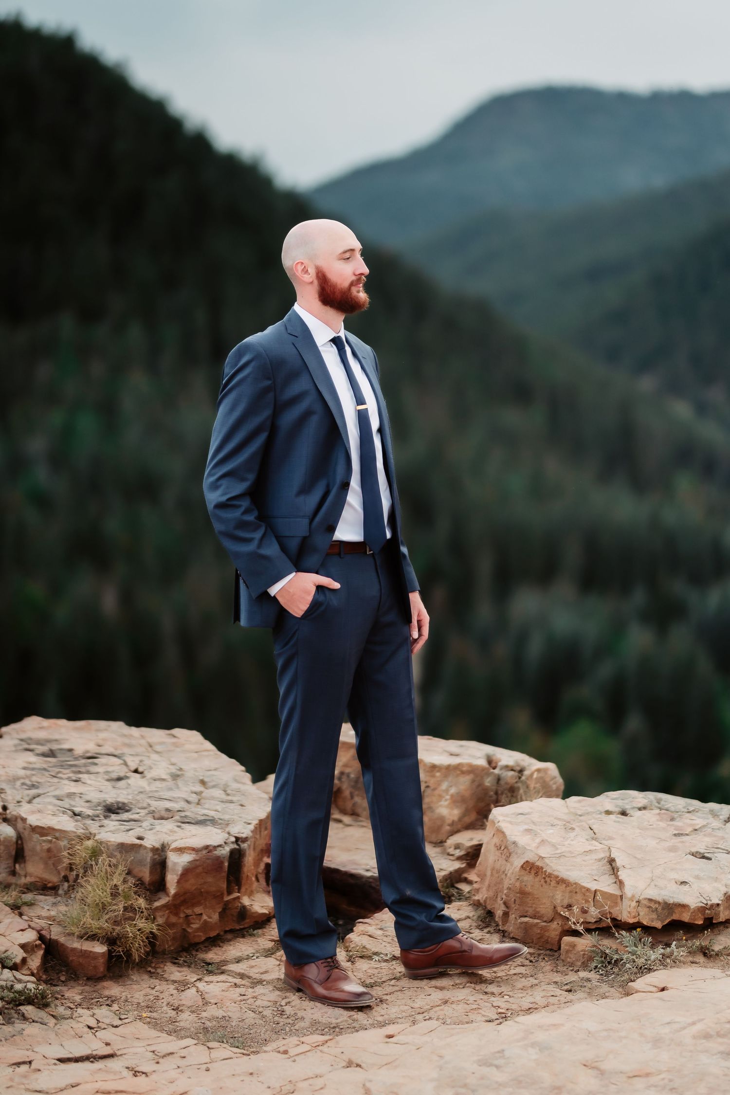 A person in a navy suit and tie stands confidently on rocky terrain with mountains and forest in the background.