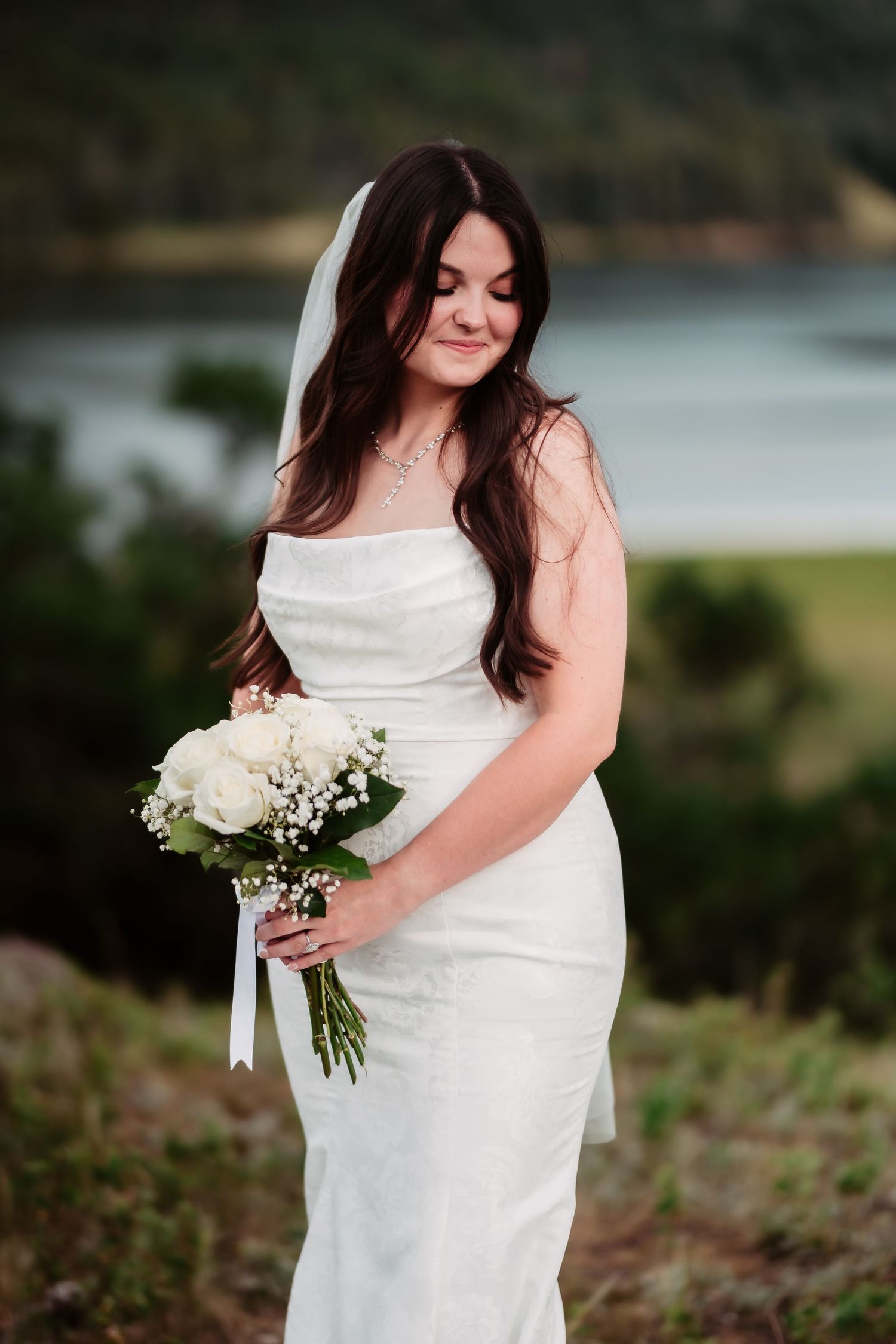 Bride in elegant white dress holding classic white bouquet against lake backdrop.