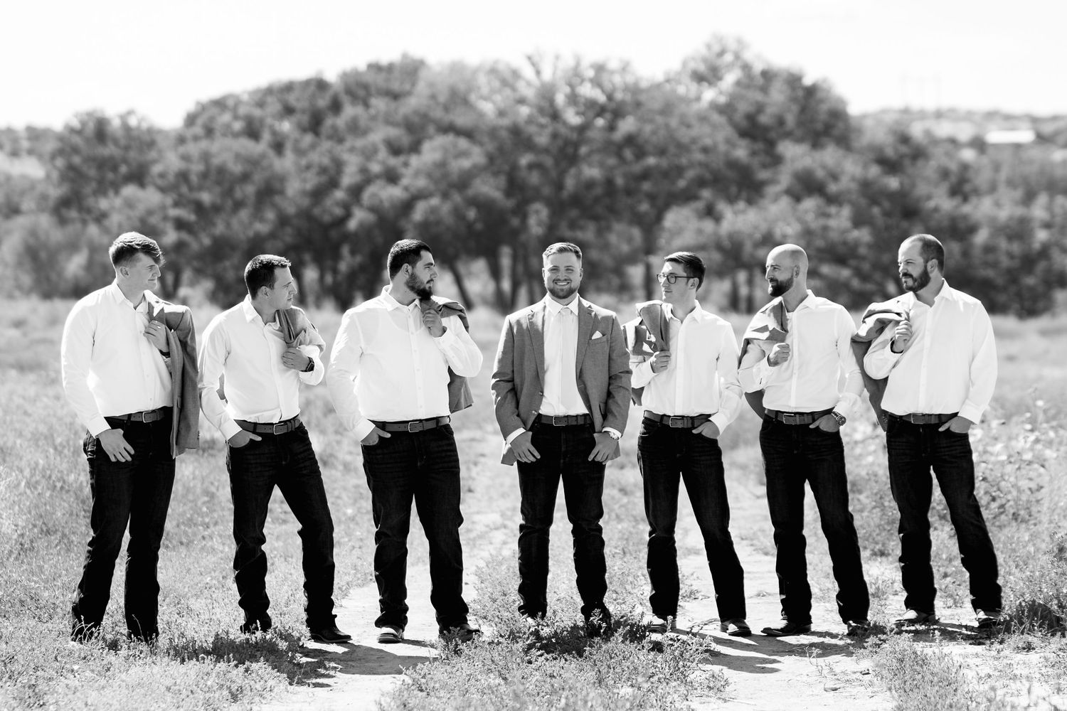 Group of groomsmen lined up in formal attire for black and white portrait in scenic field.