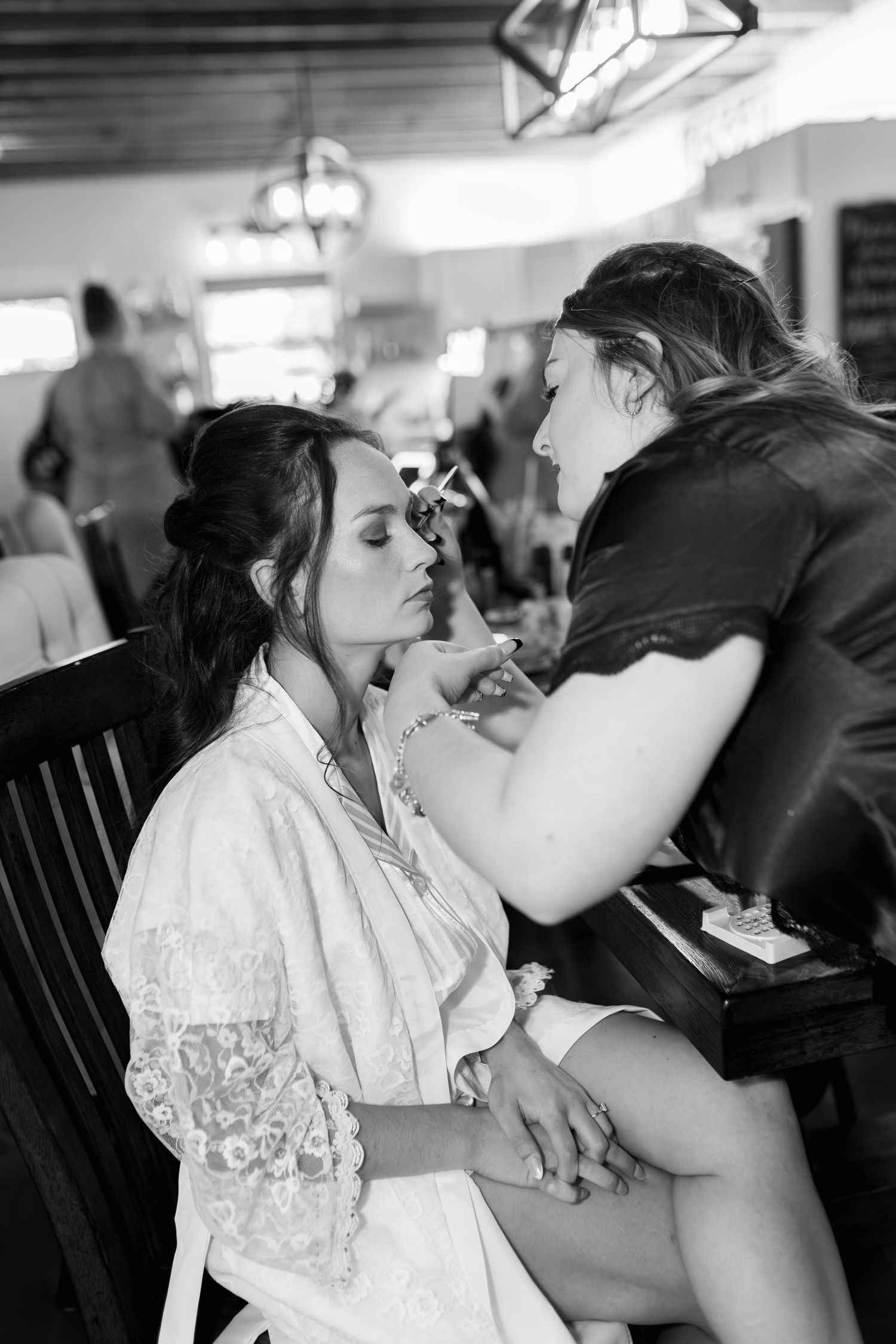 A makeup artist applies cosmetics to a person wearing a lace robe during wedding preparations.
