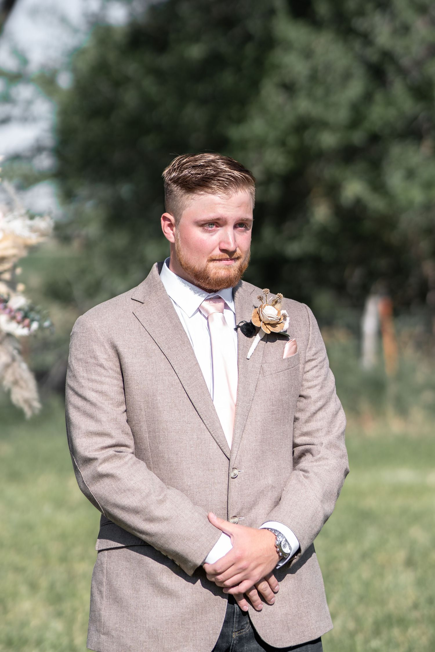 Groom in light beige suit with boutonniere stands outdoors during wedding ceremony.
