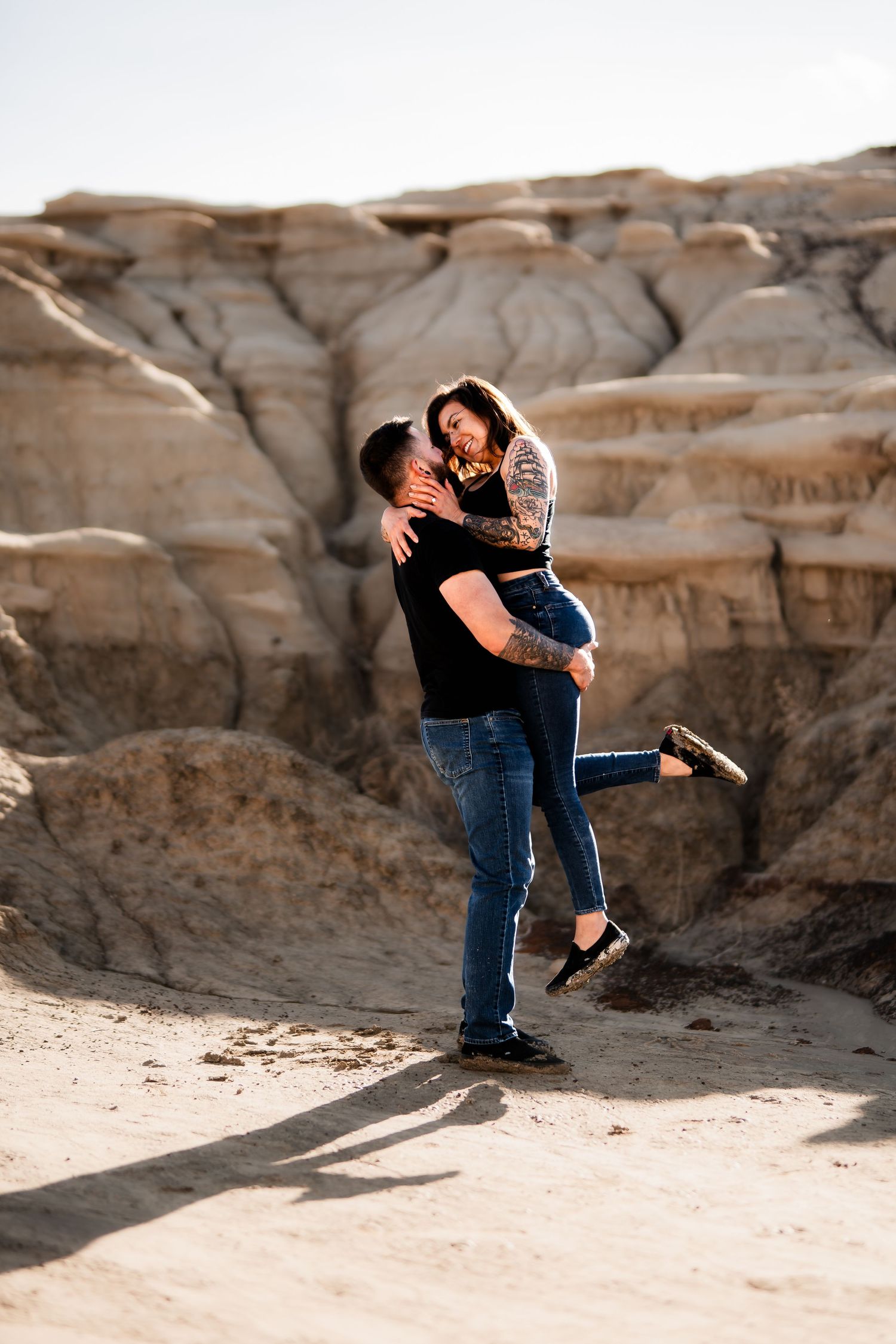 A playful romantic moment captured in front of weathered desert cliffs as one person lifts the other.