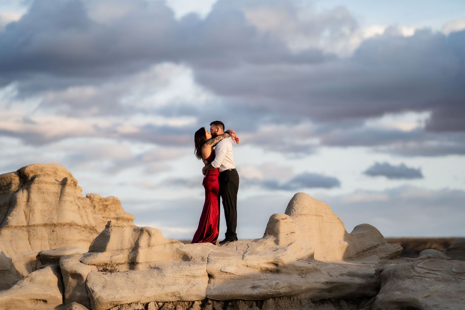 Couple embracing on desert rock formation during sunset with dramatic clouds.